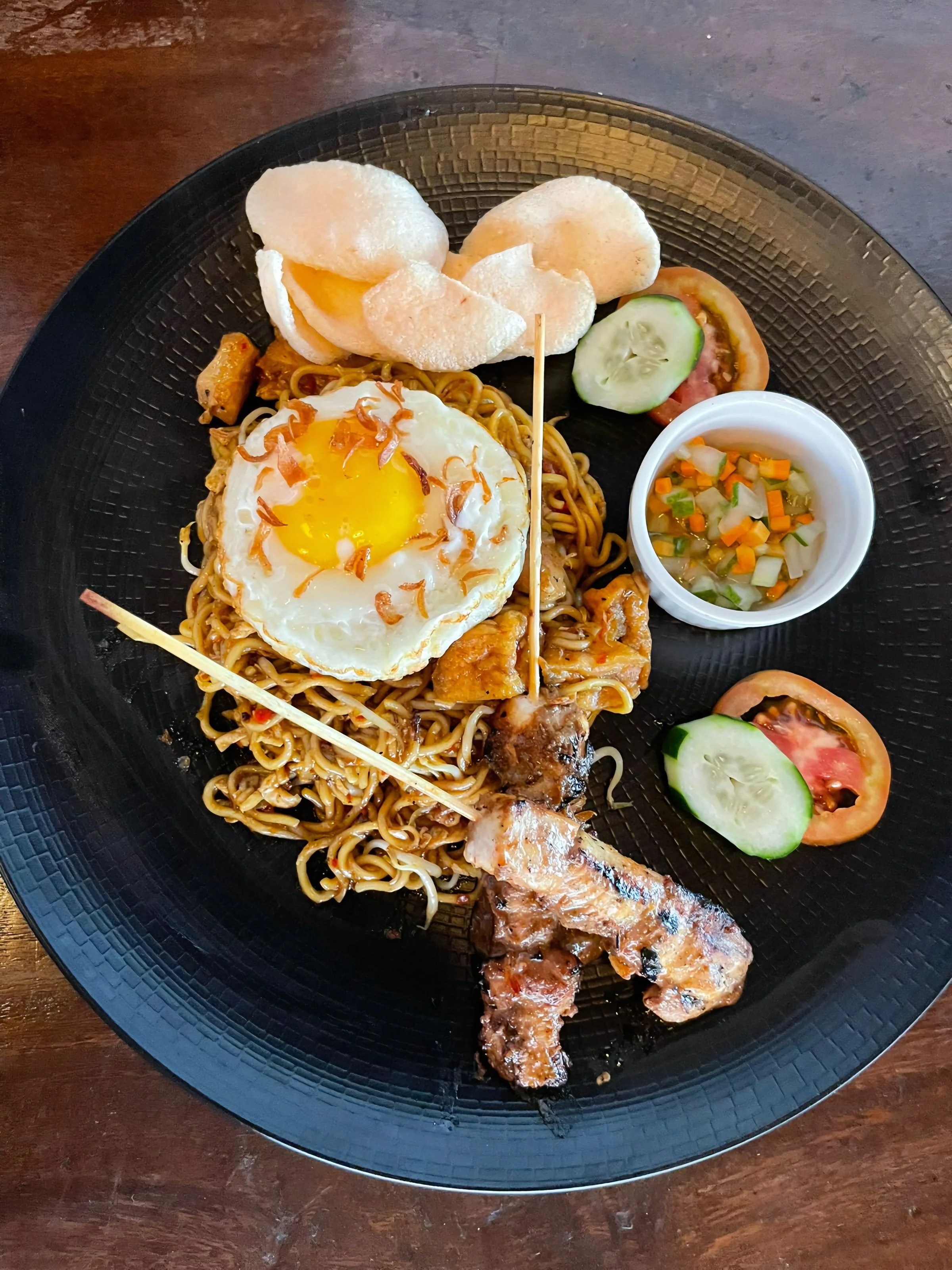 Plate of noodles with fried egg, grilled chicken leg, cucumber and tomato slices, potato chips, and a small cup of mixed chopped vegetables.