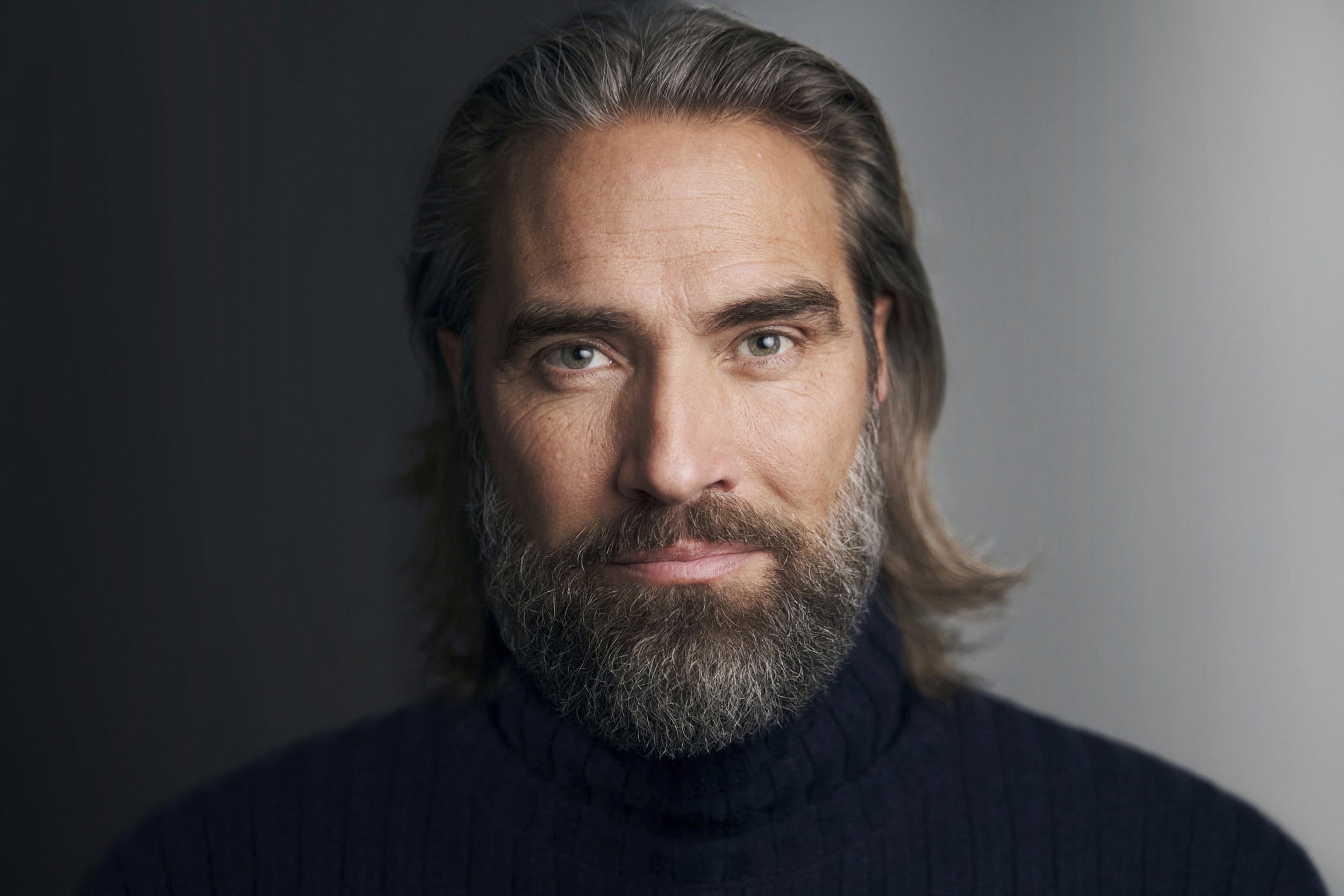 Bearded actor, close-up headshot with dramatic lighting, dark neutral background.