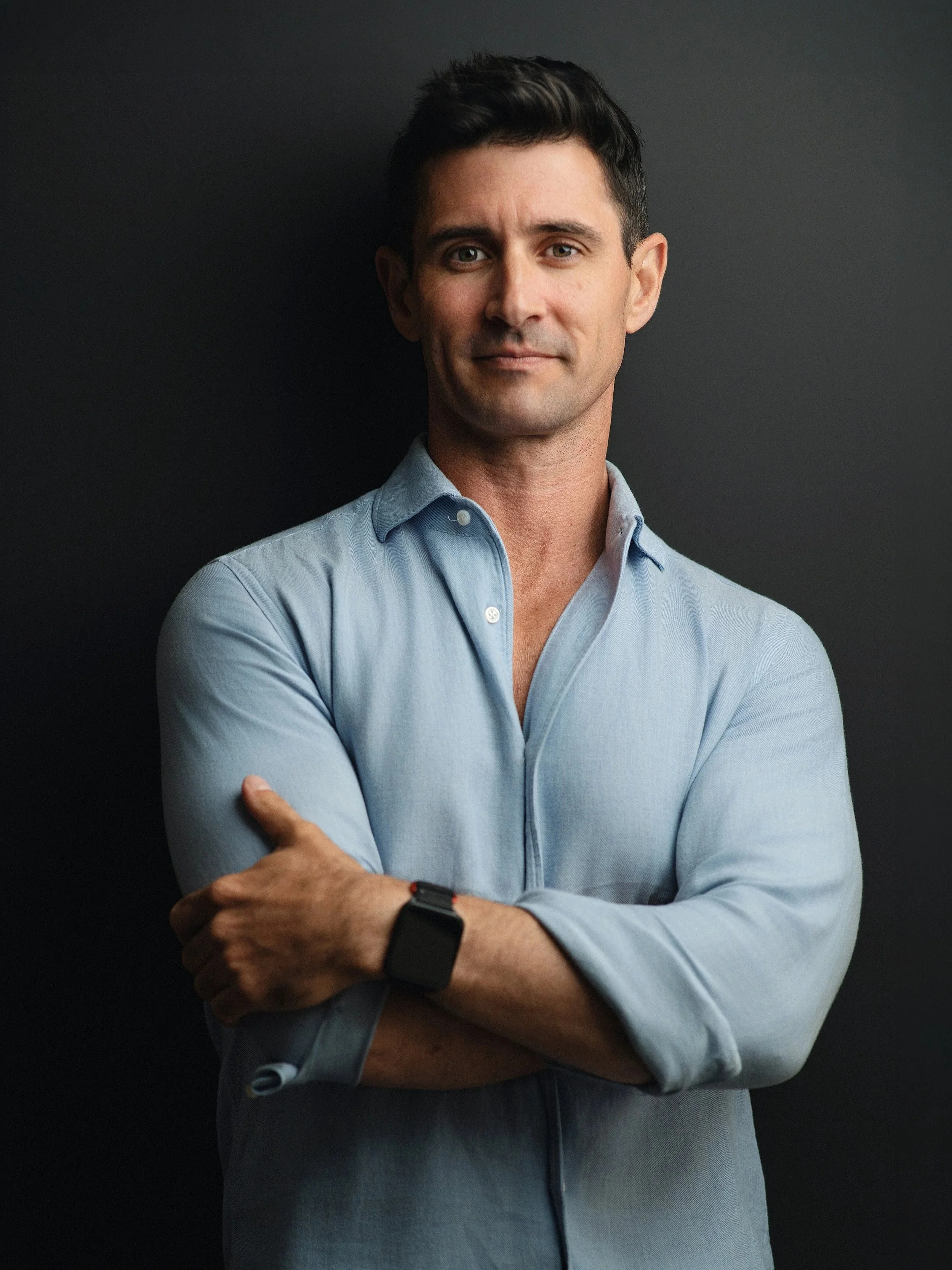 A man with dark hair, wearing a light blue button-up shirt, standing against a dark background, looking at the camera with arms crossed.