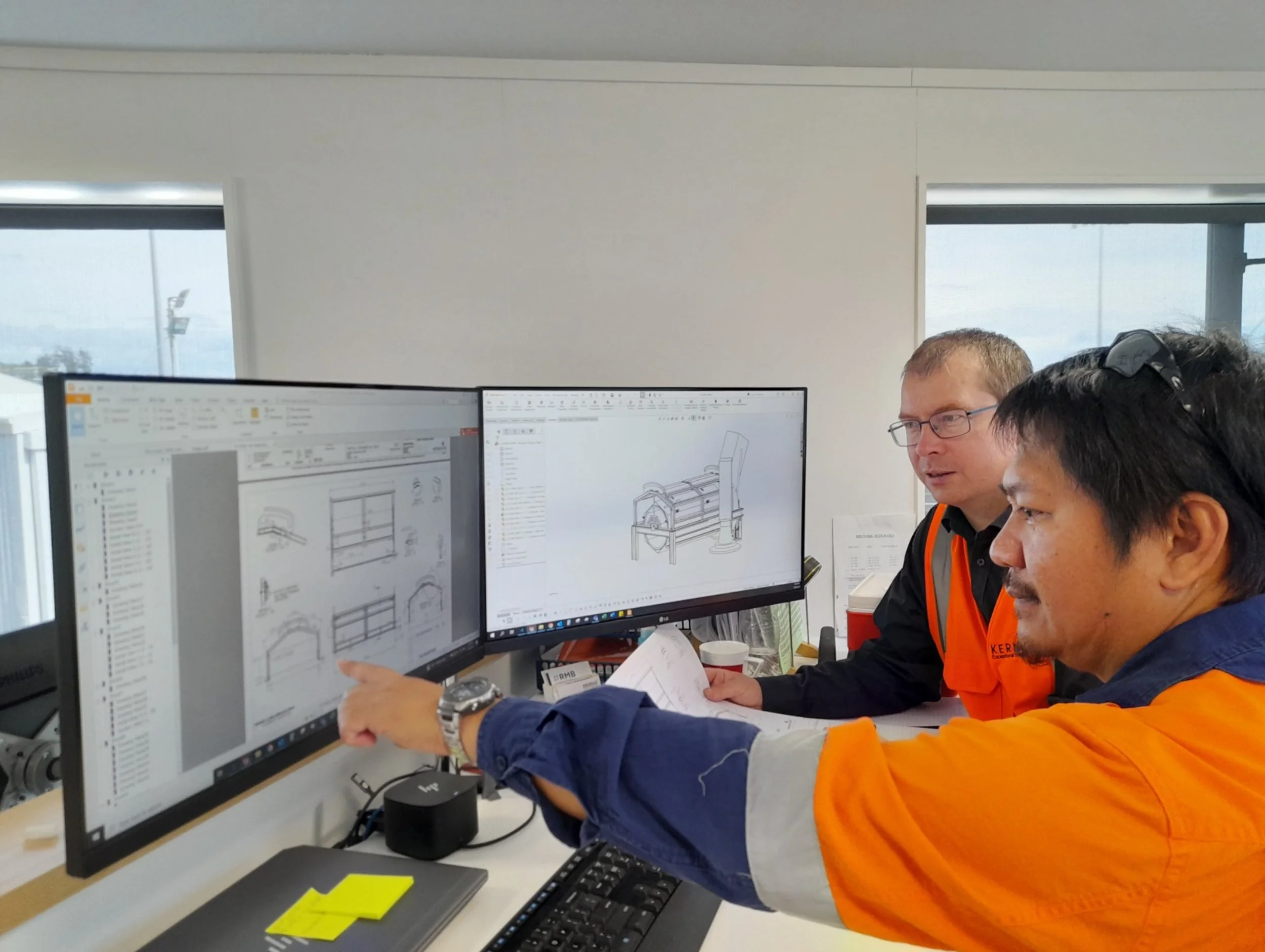 Two engineers in orange and blue safety vests examining technical drawings on dual computer monitors, discussing a machine design, in an office setting.