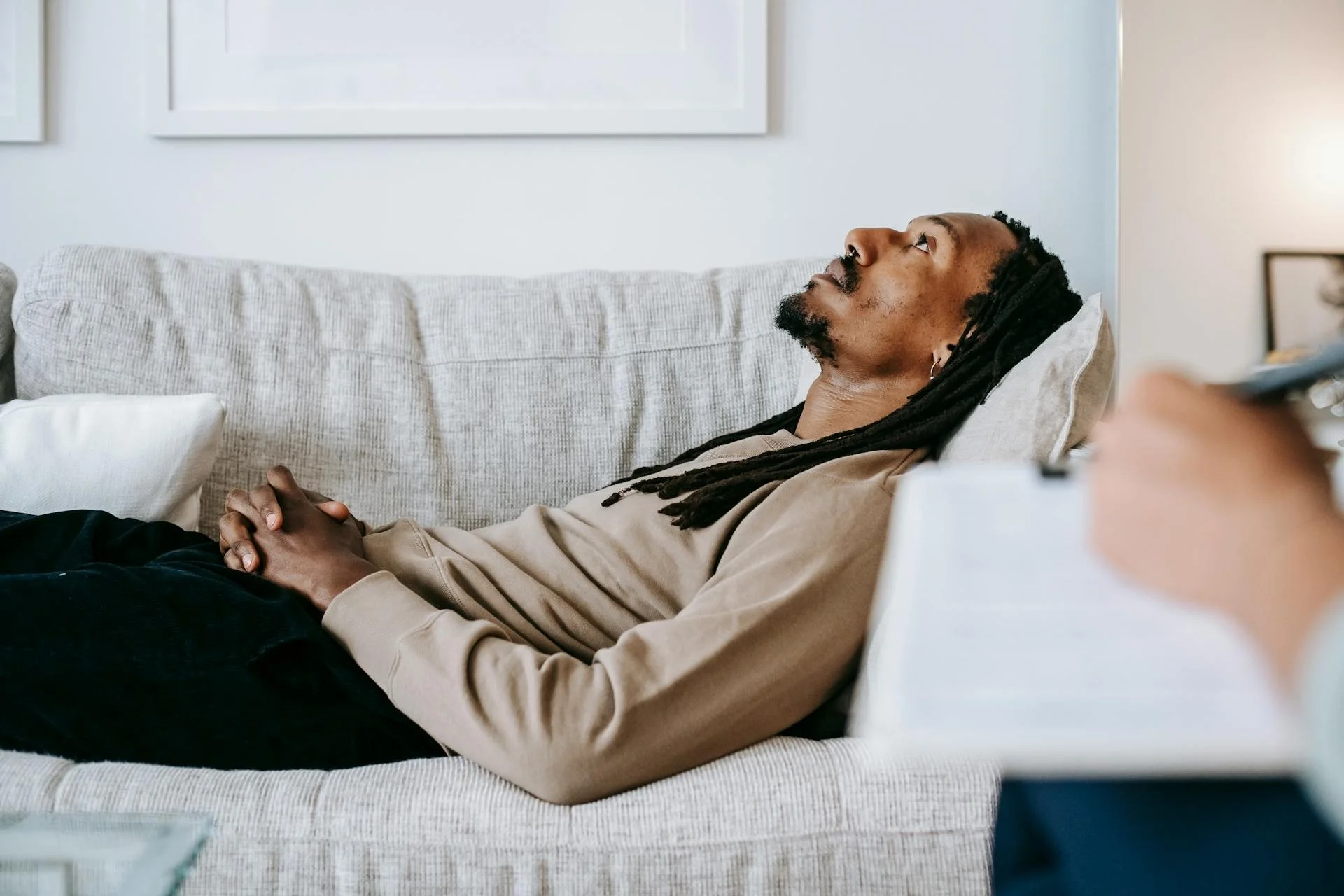 Ein Mann mit Dreadlocks liegt schlafend oder entspannt auf einem Sofa, die Hände vor der Brust verschränkt, in einem hellen Wohnzimmer.