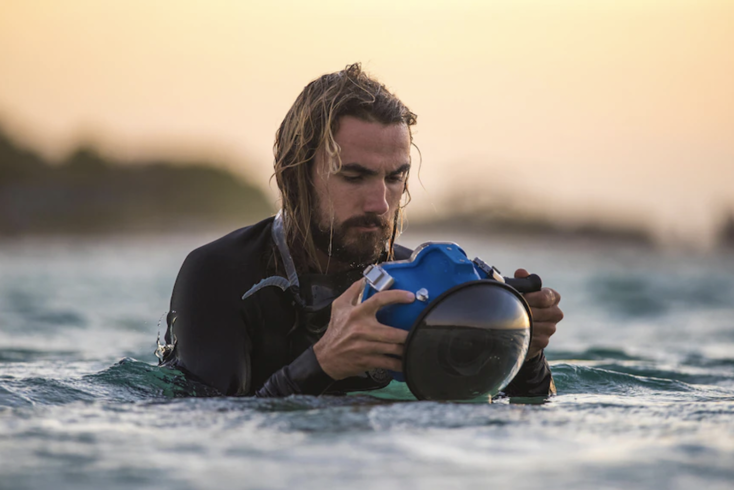 A man with long hair and a beard in water, wearing a wetsuit, examining a blue underwater camera during sunset.