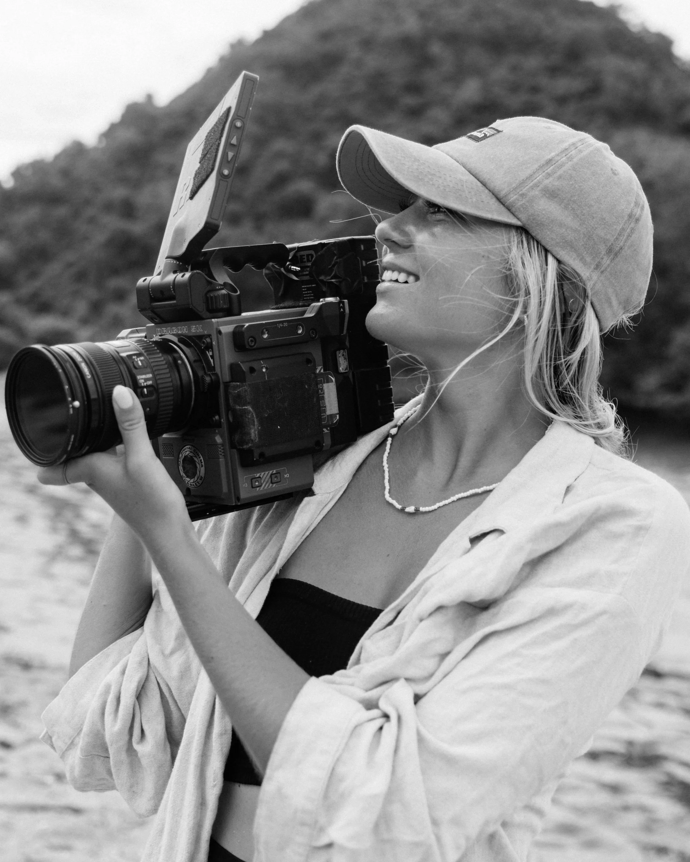 Black and white photo of a woman smiling and holding a professional video camera on her shoulder, wearing a cap and casual clothing at the beach with a hill in the background.