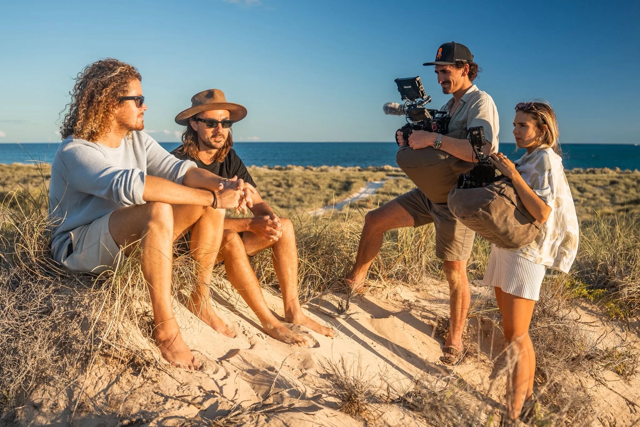 A film crew shooting a scene on a sandy beach with dunes, during sunset or late afternoon, with an ocean in the background. The crew includes two men sitting on the sand, and a man and a woman standing with professional camera equipment.