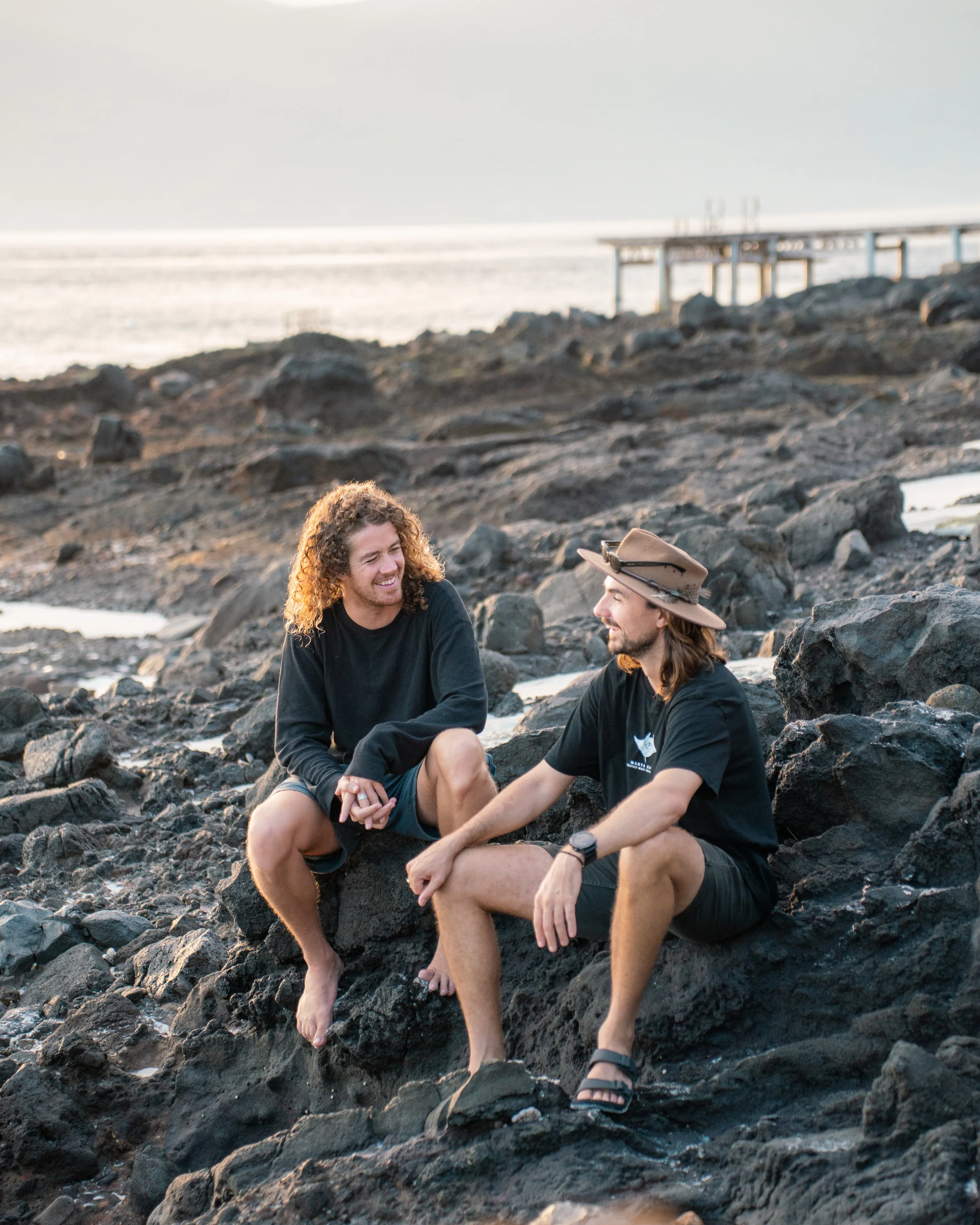 Two men sitting on rocks by the ocean, smiling at sunset, with a pier in the background.