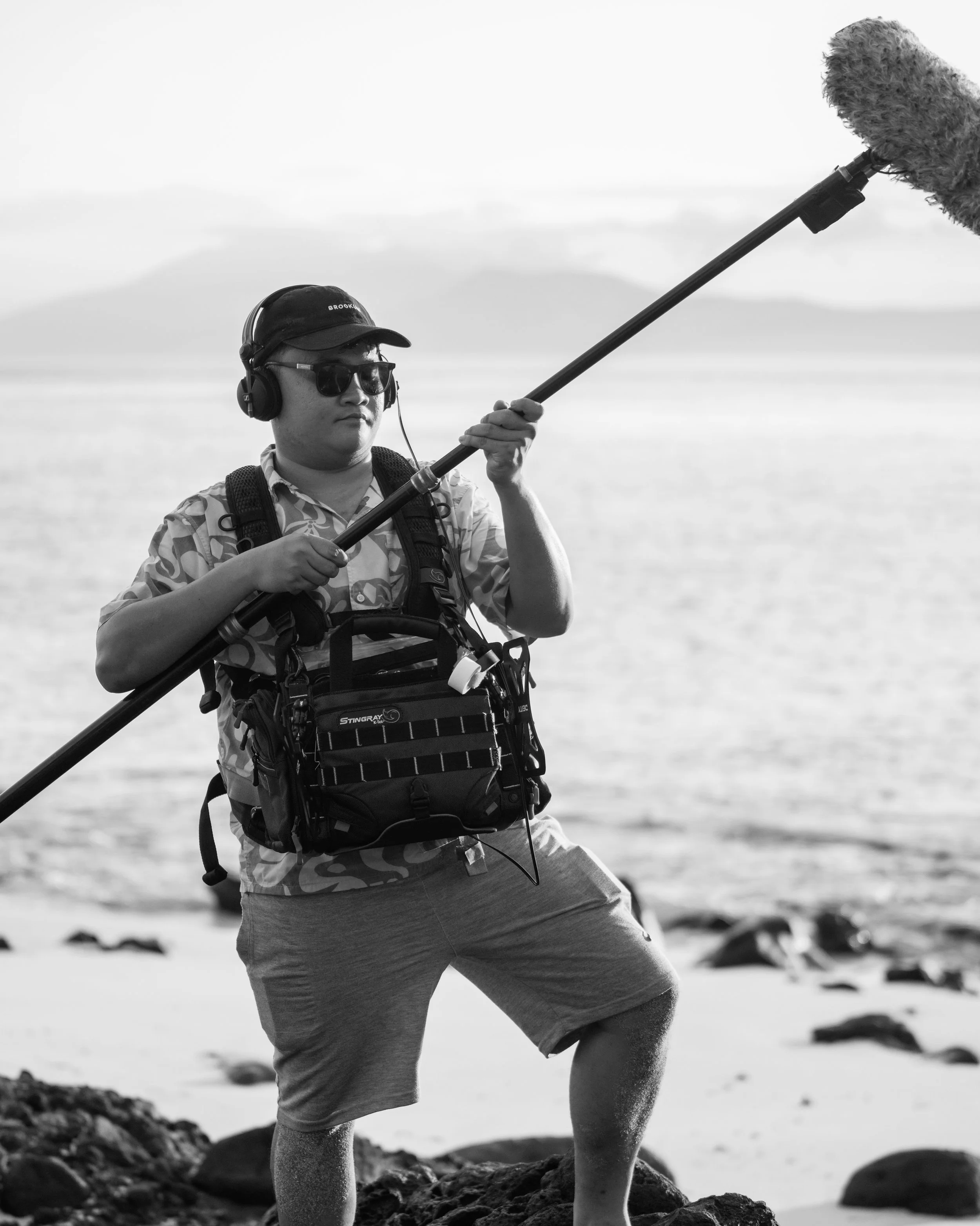 A person wearing sunglasses, a cap, and a patterned shirt holding a boom microphone at a rocky beach.