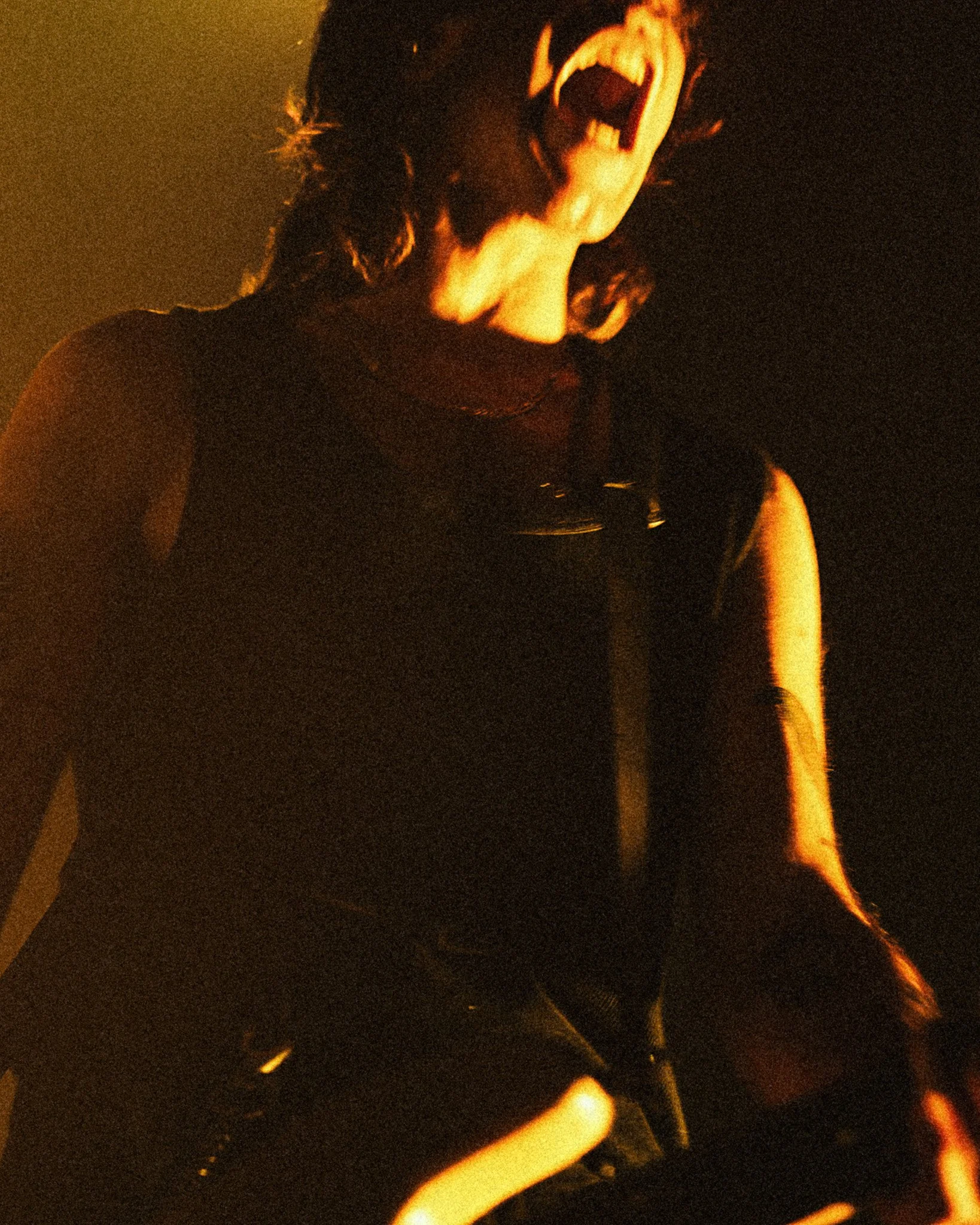 Une personne avec des cheveux longs, visible de l'épaule à la tête, criant ou chantant avec la bouche grande ouverte sous un éclairage sombre et chaud.