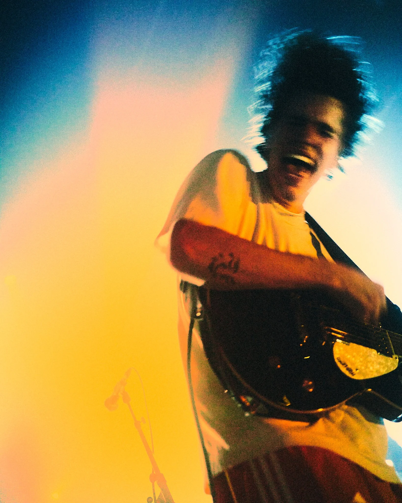 Une personne souriante avec des cheveux en ébullition joue de la guitare électrique, portant un t-shirt jaune, dans un environnement coloré et lumineux.