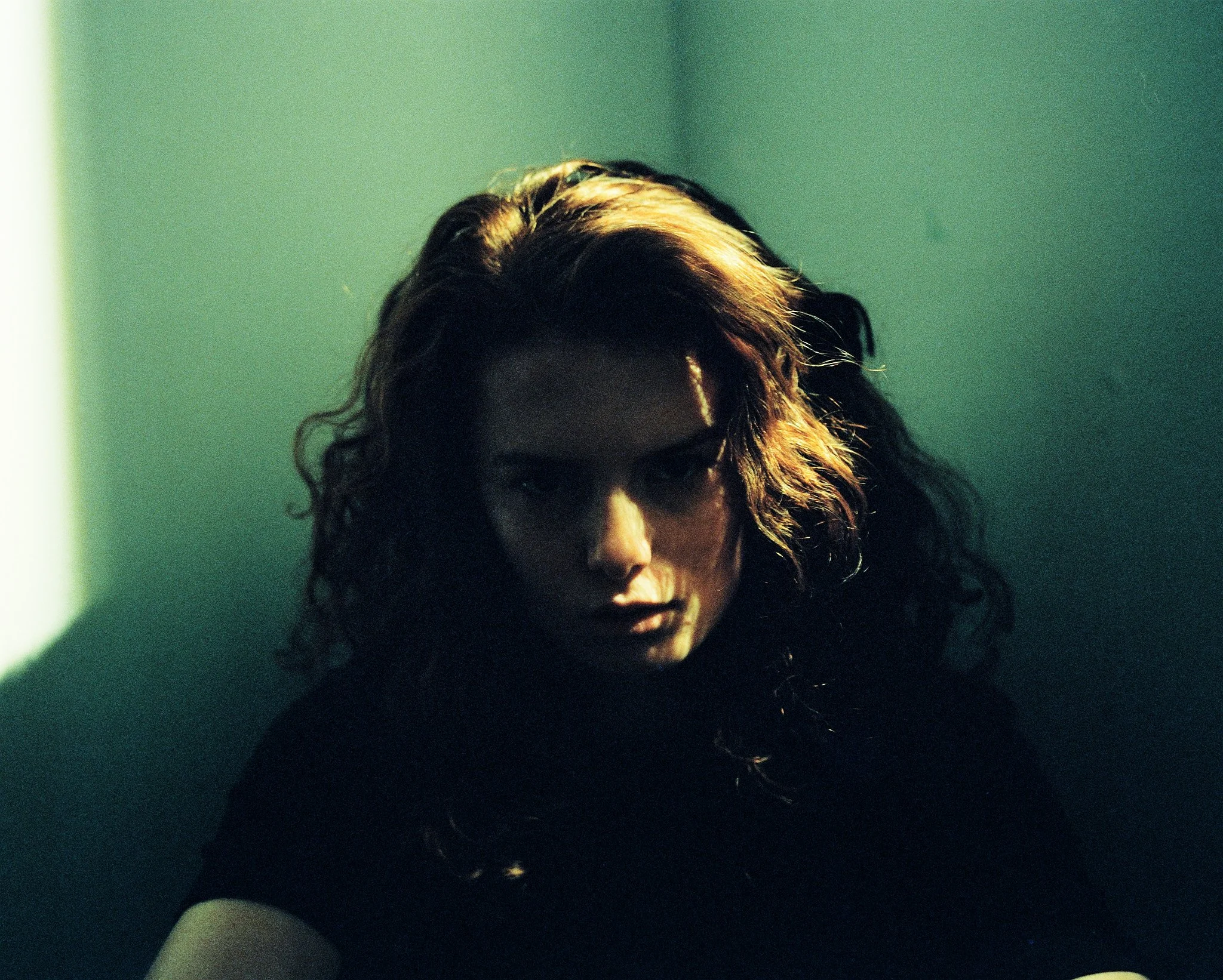 Une femme avec des cheveux bruns bouclés dans une pièce sombre, éclairée par une lumière tamisée venant de la gauche.