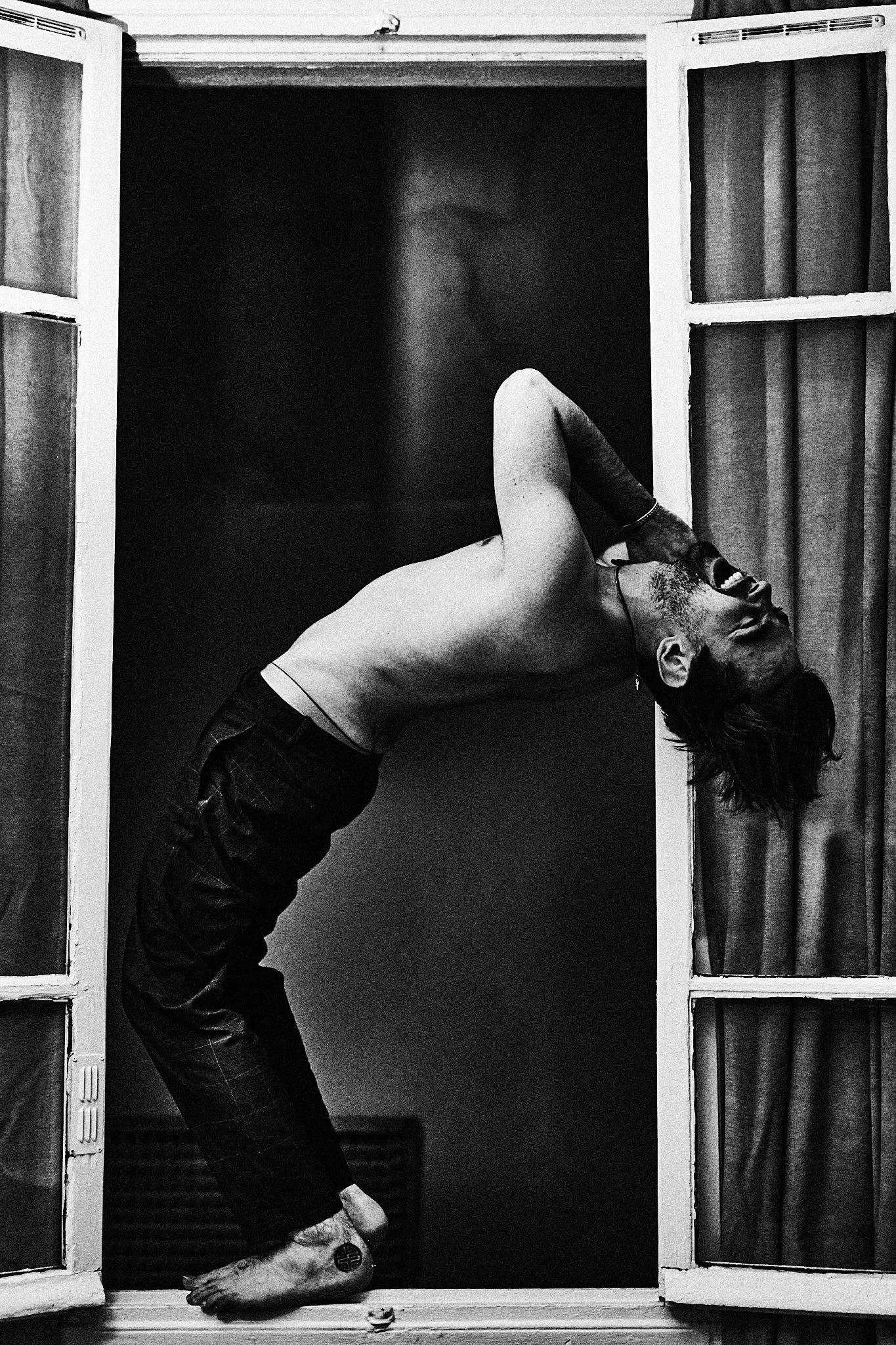 Un homme nu en équilibre, penché en arrière, dans un cadre de porte avec rideaux, en noir et blanc.