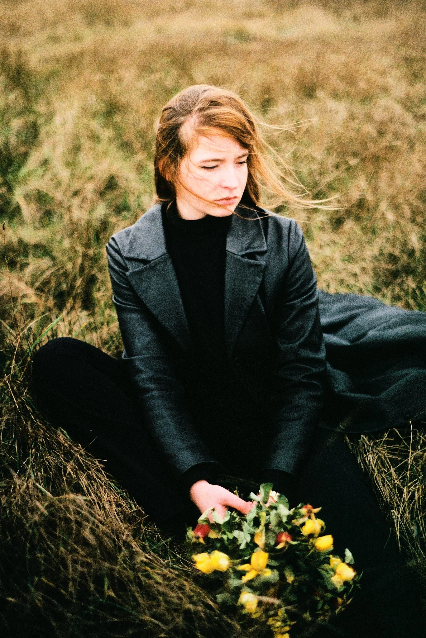 Une jeune femme aux cheveux bruns et bouclés, vêtue d'une veste en cuir noire, assise dans une prairie, tenant un bouquet de fleurs jaunes et rouges. Elle a l'air pensive, regardant vers le bas.