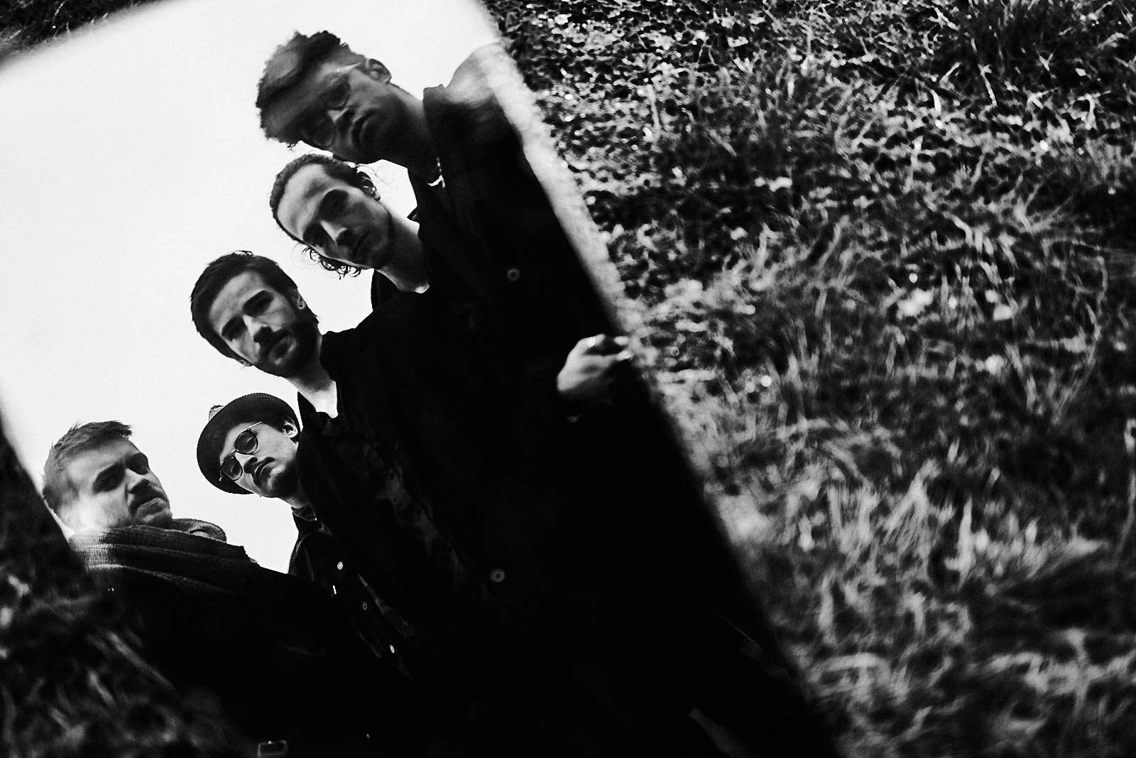 Cinq jeunes hommes posant à l'extérieur, photographiés en noir et blanc, certains portant des lunettes et des chapeaux, regardant vers l'appareil photographique, devant un fond de nature avec de l'herbe.