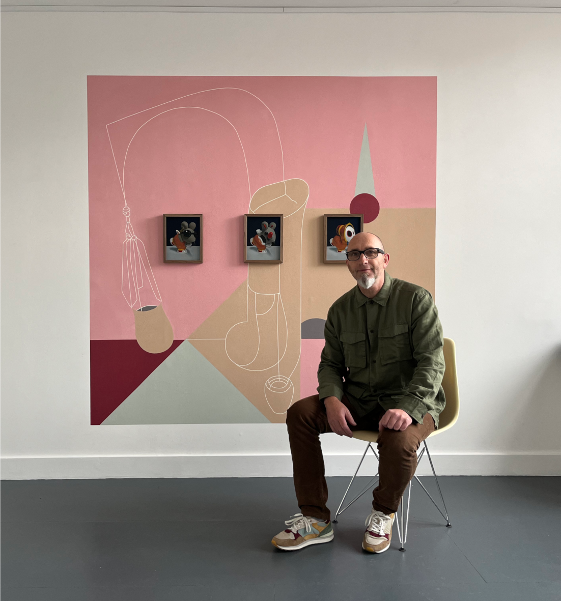 An  artist glasses, a beard, and a goatee sitting on a yellow chair in front of a pink and neutral abstract art mural with framed paintings of image+text generated toys.