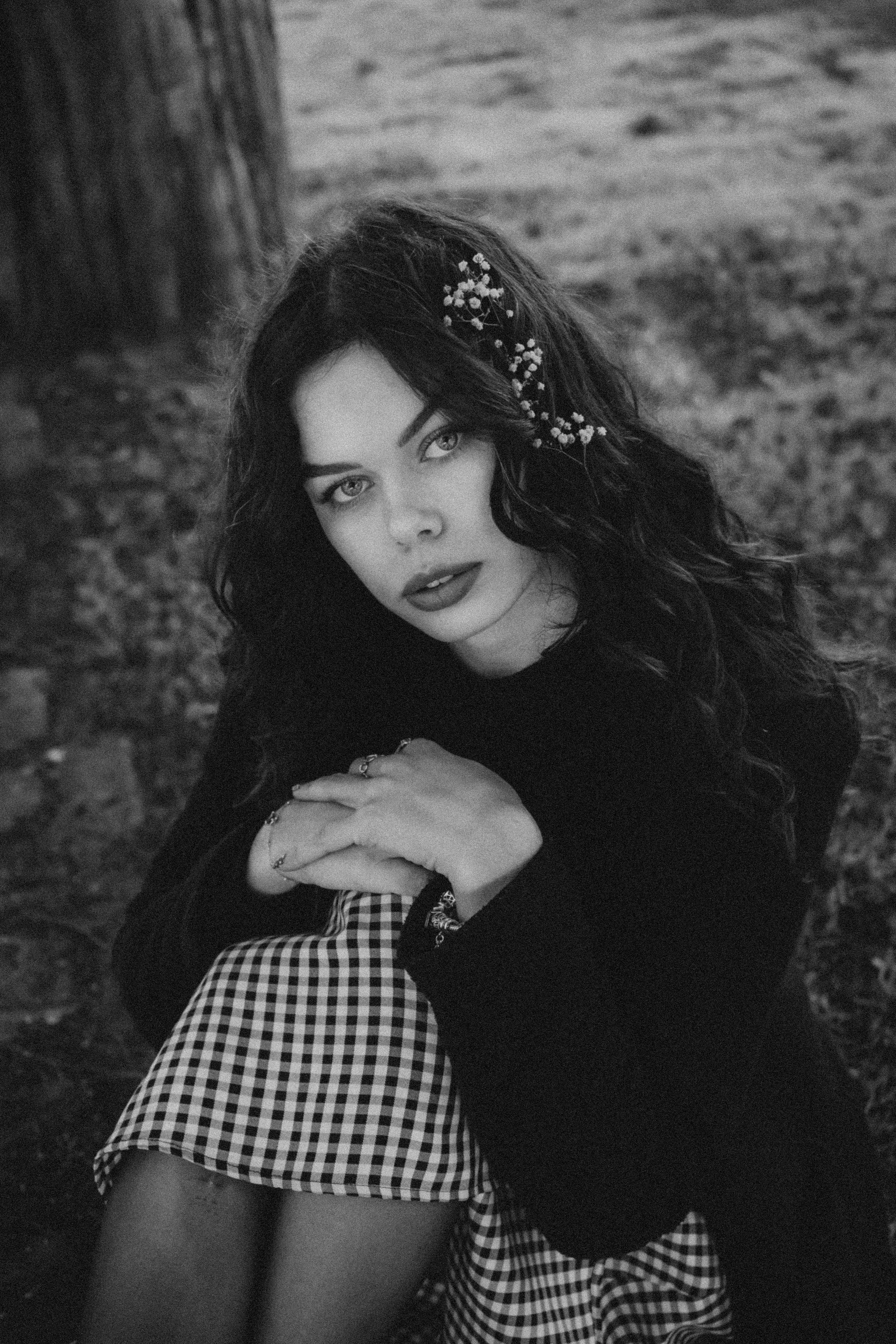 Une jeune femme aux cheveux bouclés, portant une robe à carreaux, assise près d'un arbre, dans un environnement naturel, en noir et blanc.
