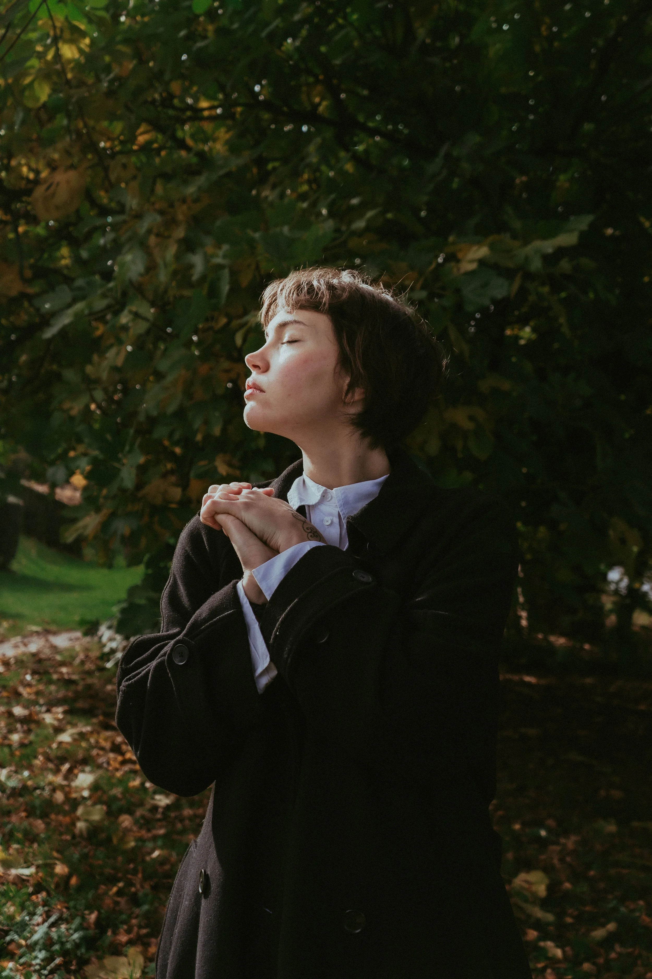 Une jeune femme aux cheveux courts, en vêtement noir avec une chemise blanche, se tient en plein air dans un parc, les yeux fermés, les mains jointes devant sa poitrine, sous un arbre aux feuilles d'automne.