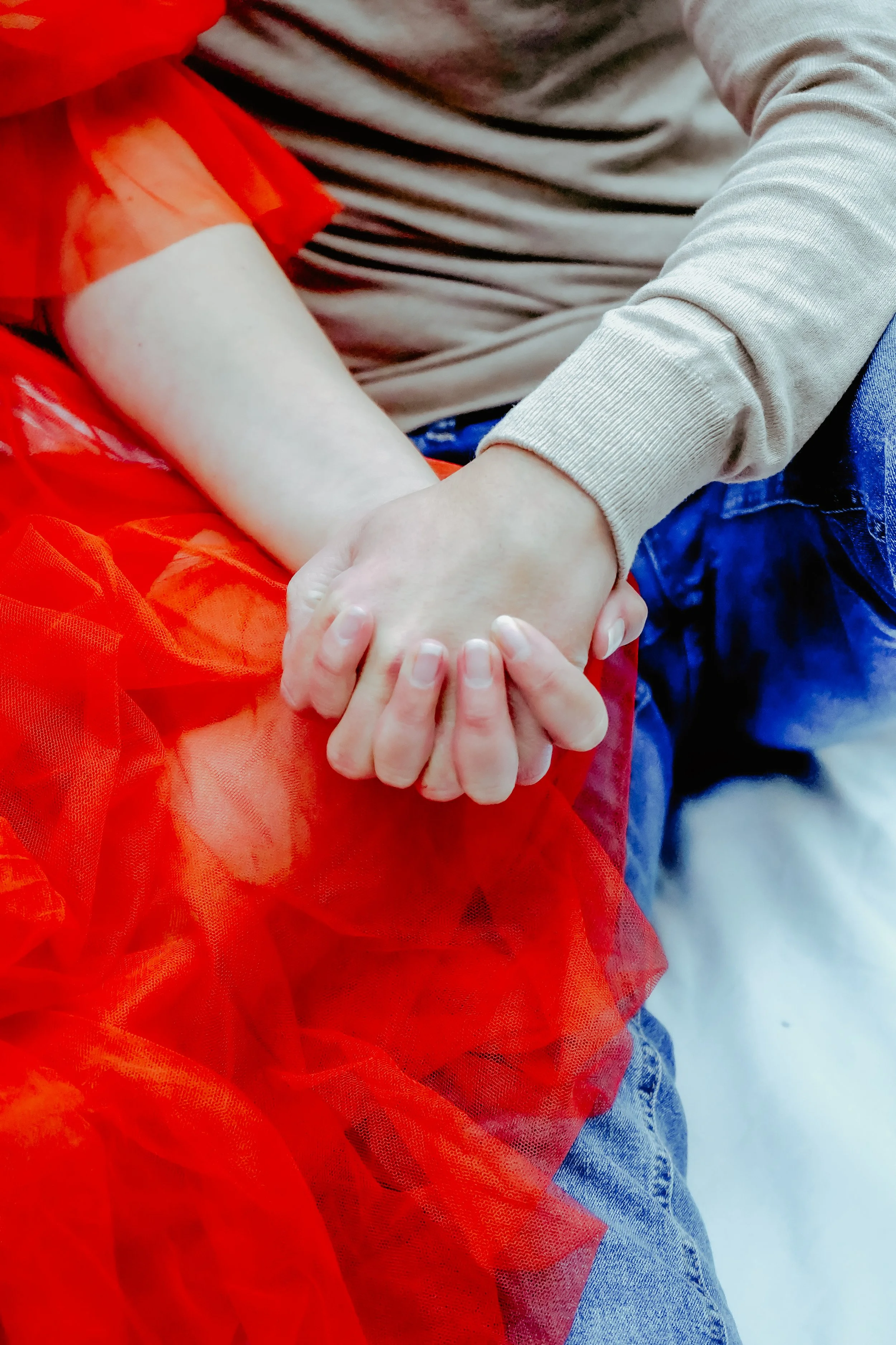 Deux personnes tiennent la main l'une de l'autre, avec le tissu rouge vif autour de leur bras. La main de gauche appartient à une personne portant une manche beige, et la main de droite appartient à une personne portant un pantalon en jean bleu.