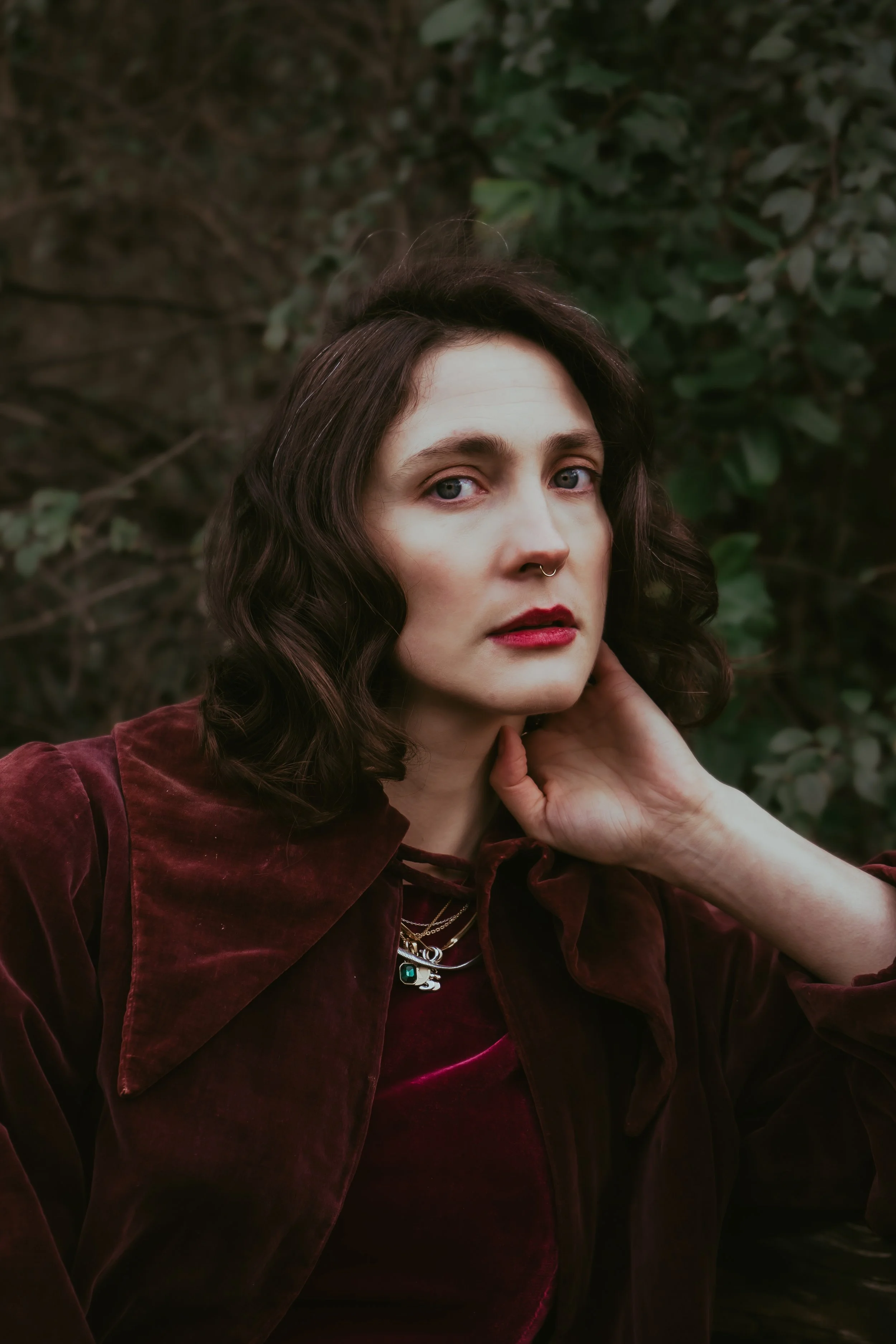Une femme aux cheveux foncés, portant un manteau en velours bordeaux, posant devant un fond de feuillages verts, avec des bijoux autour du cou.