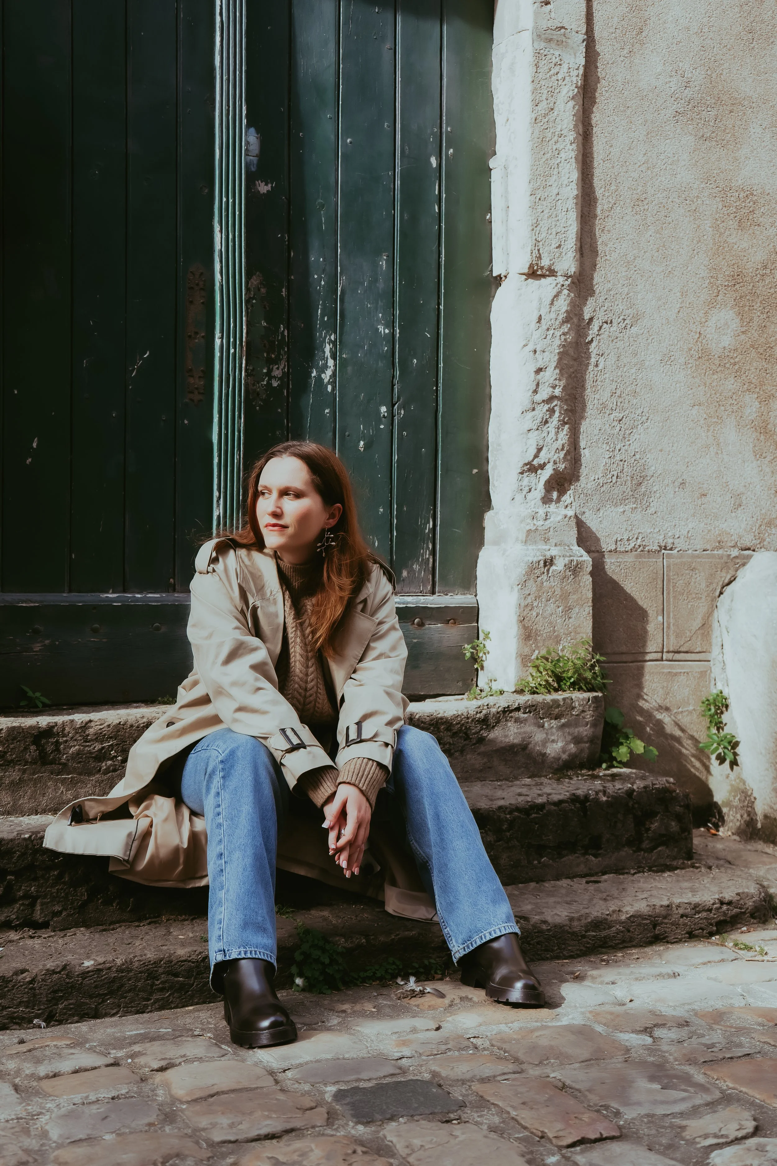 Une femme assise sur des marches en pierre devant une porte en bois vert et un mur en pierre. Elle porte un trench beige, un pull marron, un jean bleu et des bottes noires.