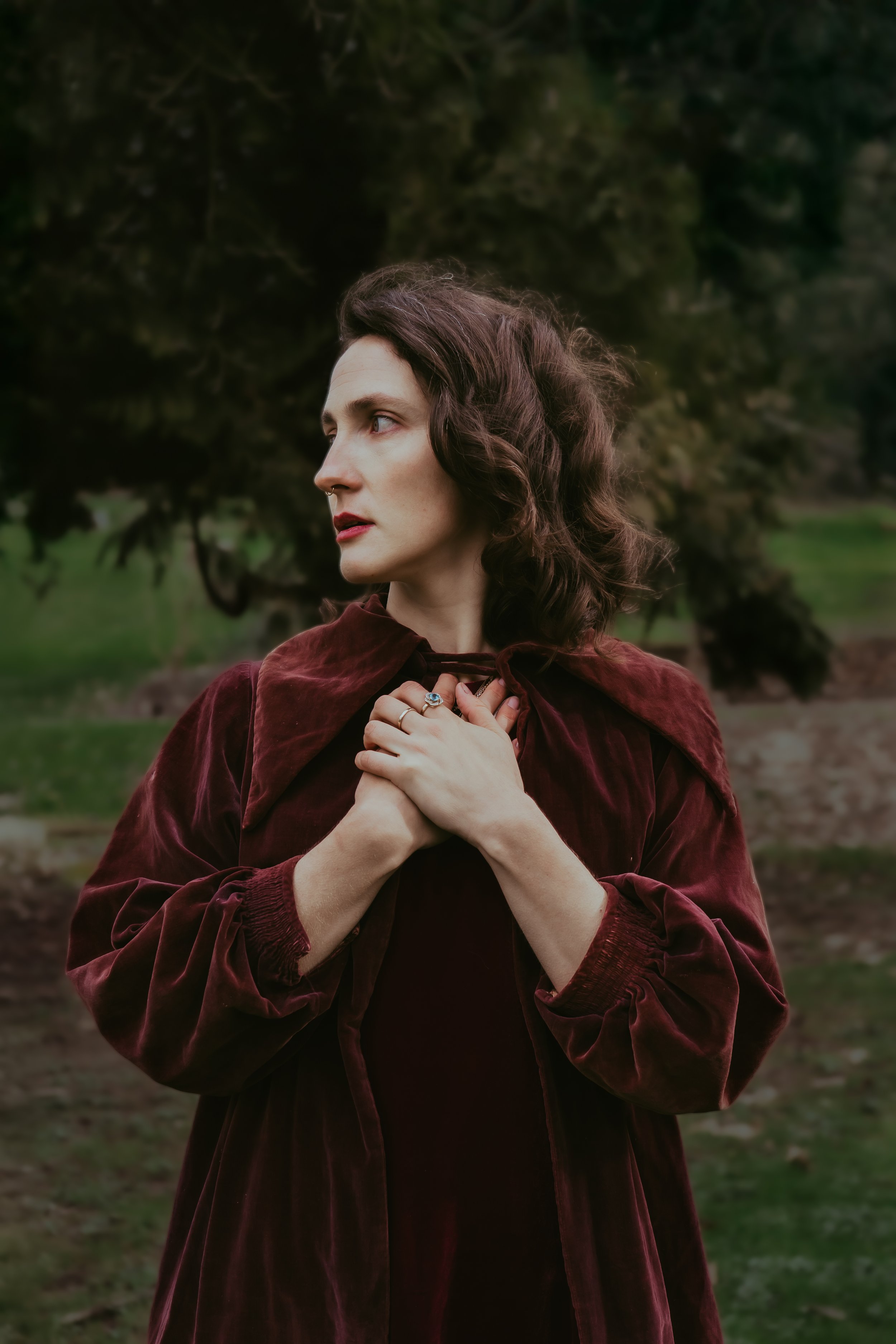 Femme avec cheveux ondulés et robe en velours bordeaux, mains croisées sur la poitrine, dans un parc avec des arbres en arrière-plan.