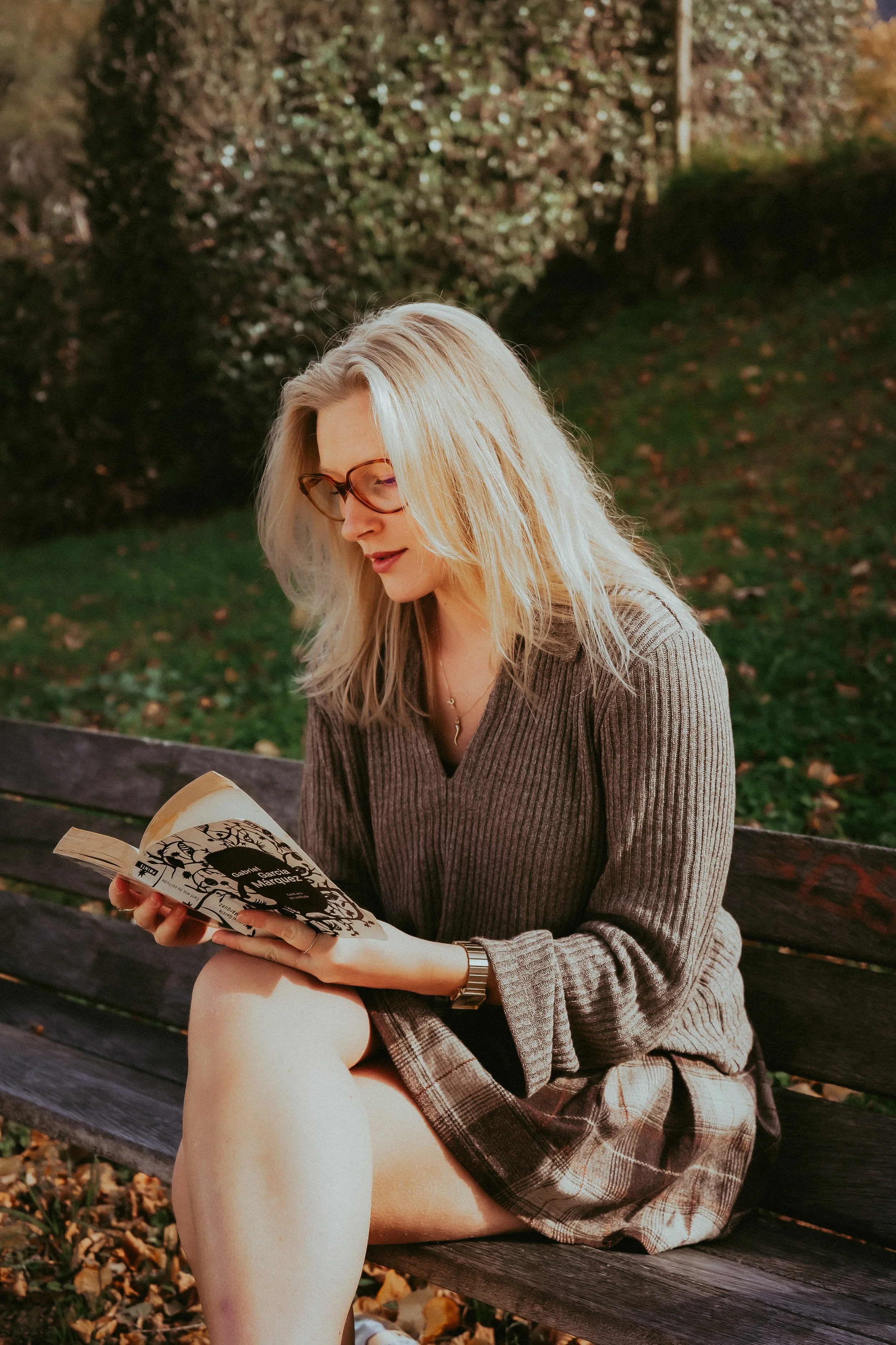Une femme aux cheveux blonds lisant un livre assise sur un banc dans un parc en automne.