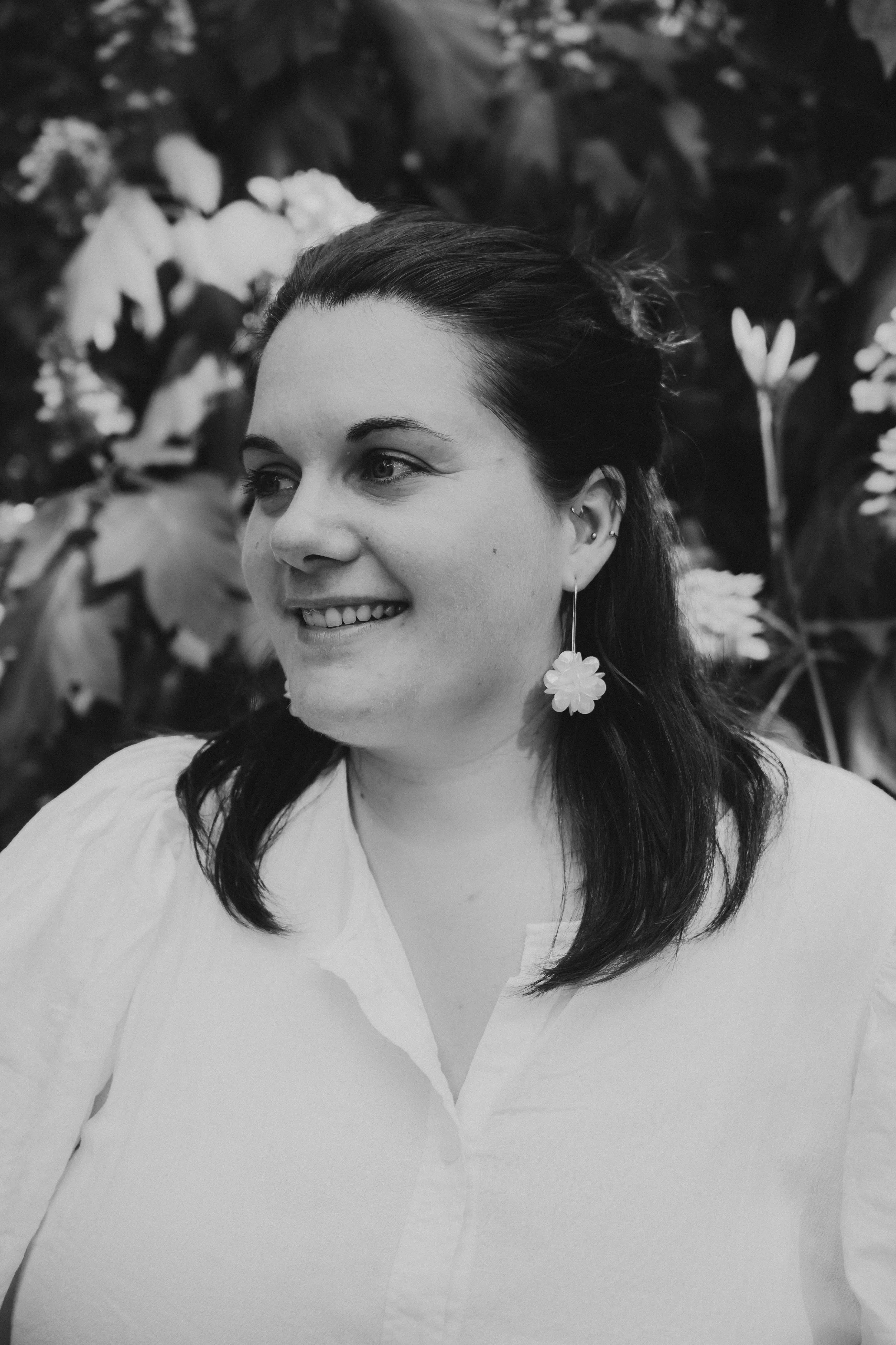 Portrait en noir et blanc d'une femme souriante avec de longs cheveux noirs, portant une paire de boucles d'oreilles en forme de fleur, en arrière-plan de feuillage.