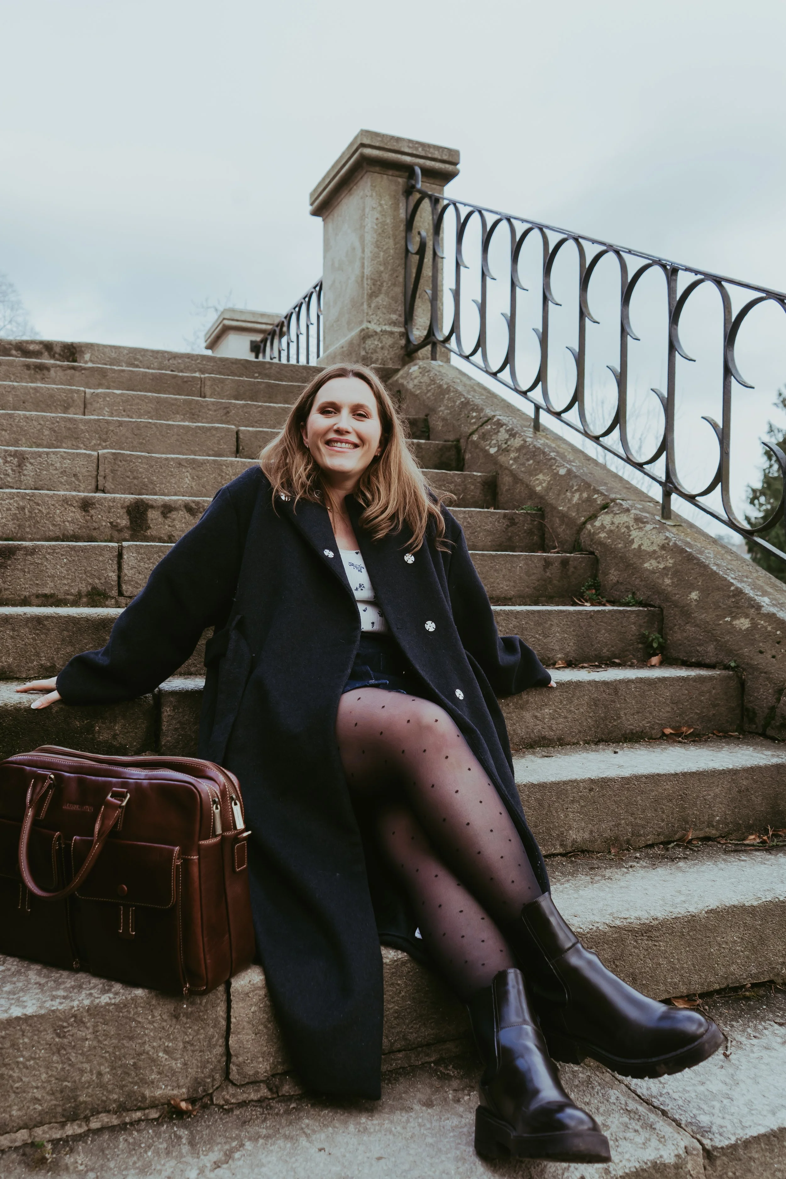 Une femme souriante assise sur des marches en pierre avec une valise en cuir à côté, portant un manteau noir, des collants à pois et des bottines en cuir, devant une rambarde en métal et un ciel nuageux.