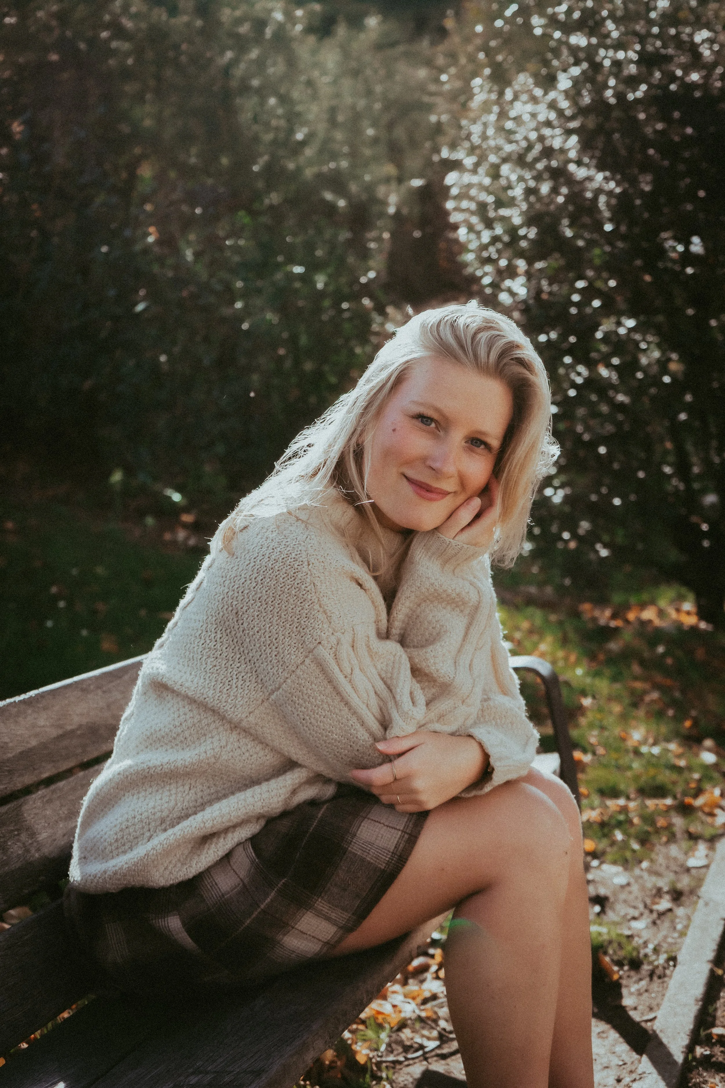 Une jeune femme blonde souriante assise sur un banc en extérieur, portant un pull beige et une jupe à carreaux, avec des arbres en arrière-plan et la lumière du soleil filtre à travers les feuilles.