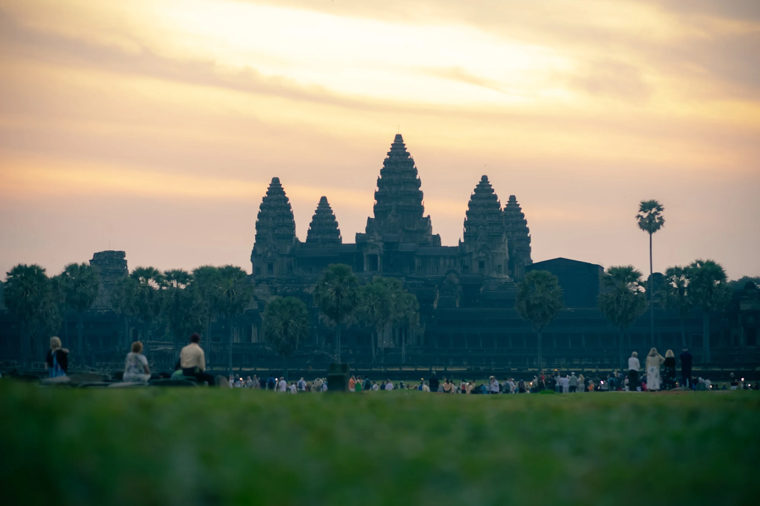 Silhouette of Angkor Wat temple in Siem Reap, Cambodia at sunset with people and palm trees in the foreground.