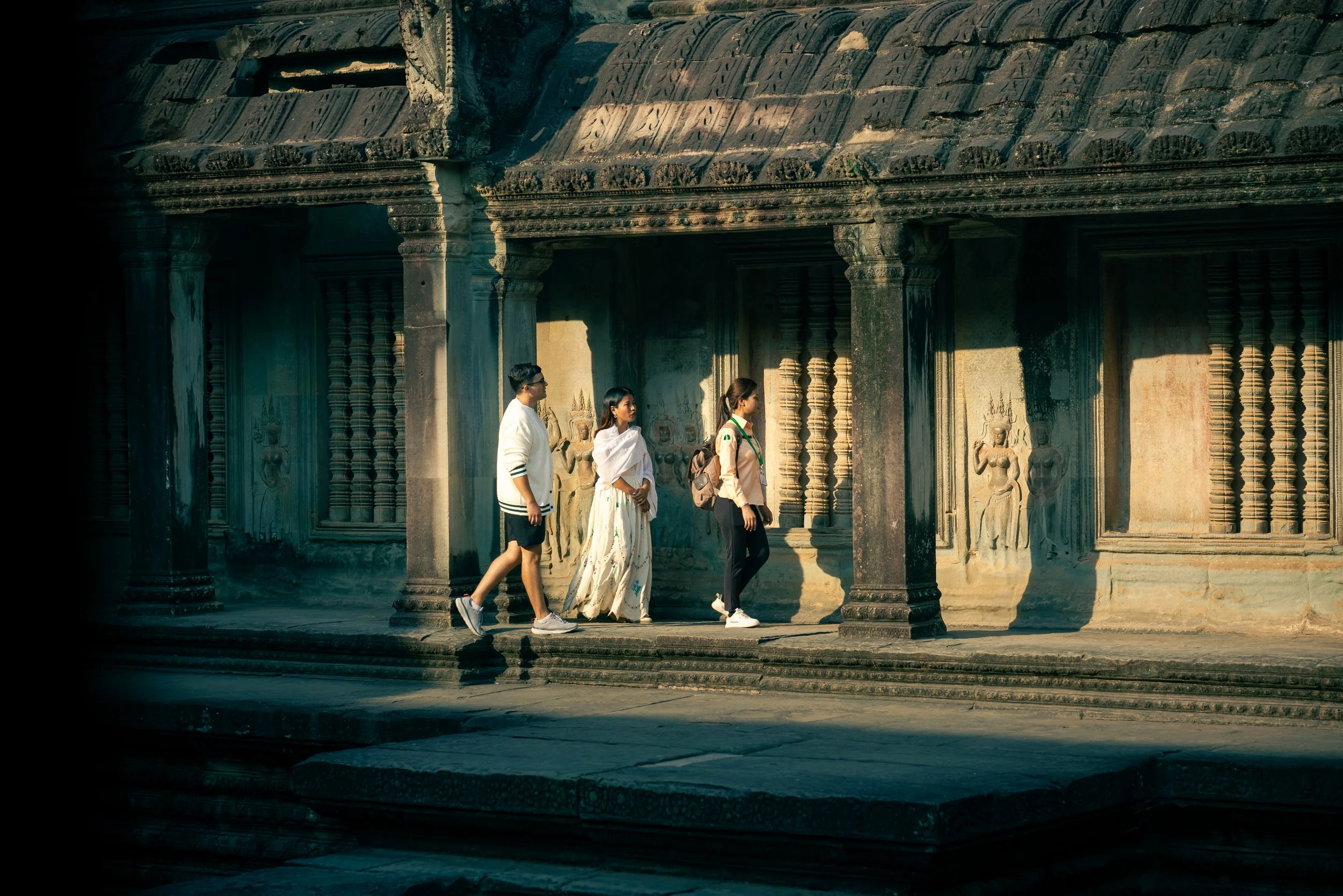 Three people walking past an ancient stone building Angkor Wat with columns and carved reliefs in Siem Reap, Cambodia.