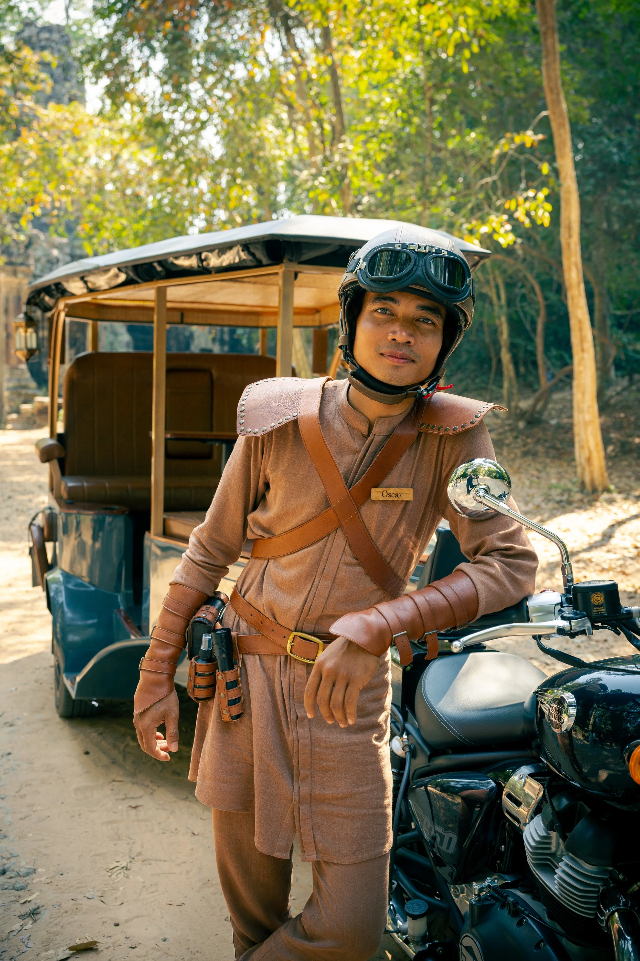 A driver wearing a helmet and leather outfit standing next to a motorcycle in Angkor forest. Behind them is a tuk-tuk with forest trees in the background in Siem Reap, Cambodia.
