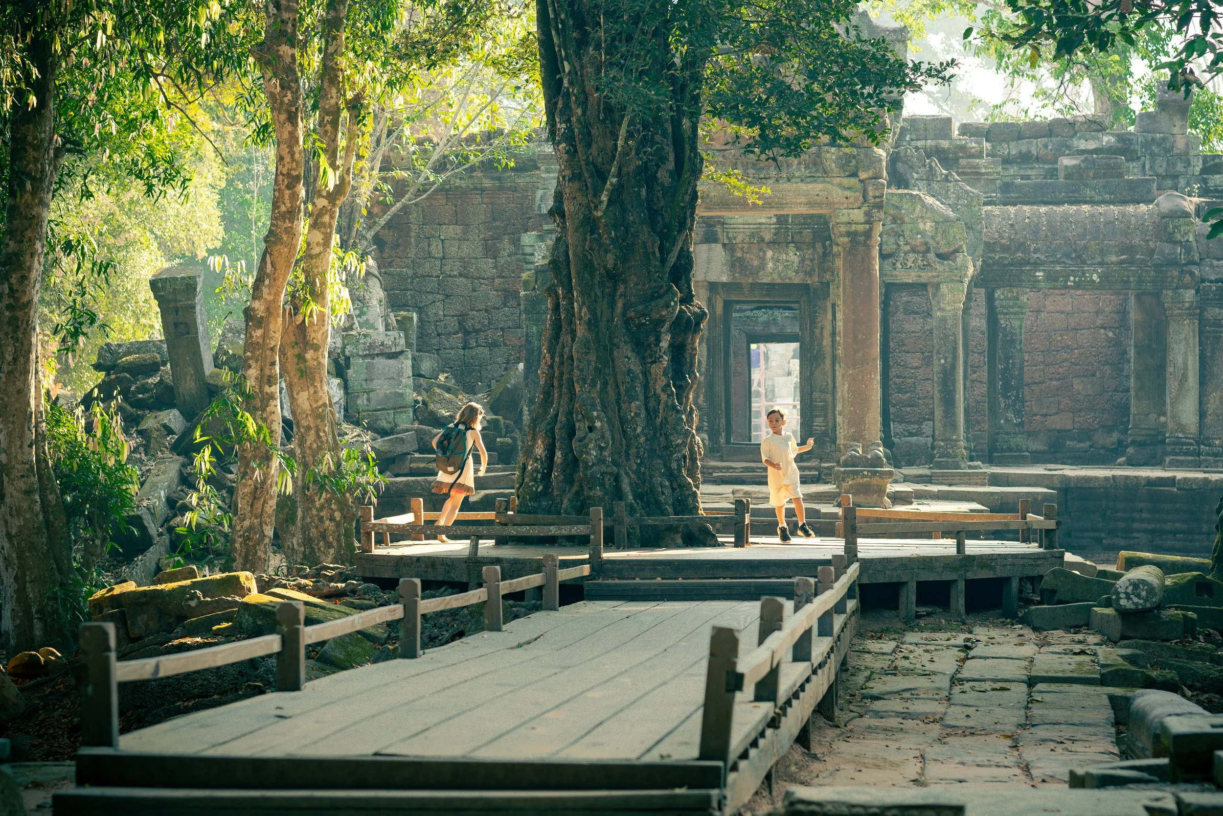 Two people walking along a wooden path through ancient temple Ta Prohm ruins surrounded by trees and stone structures in Siem Reap, Cambodia.