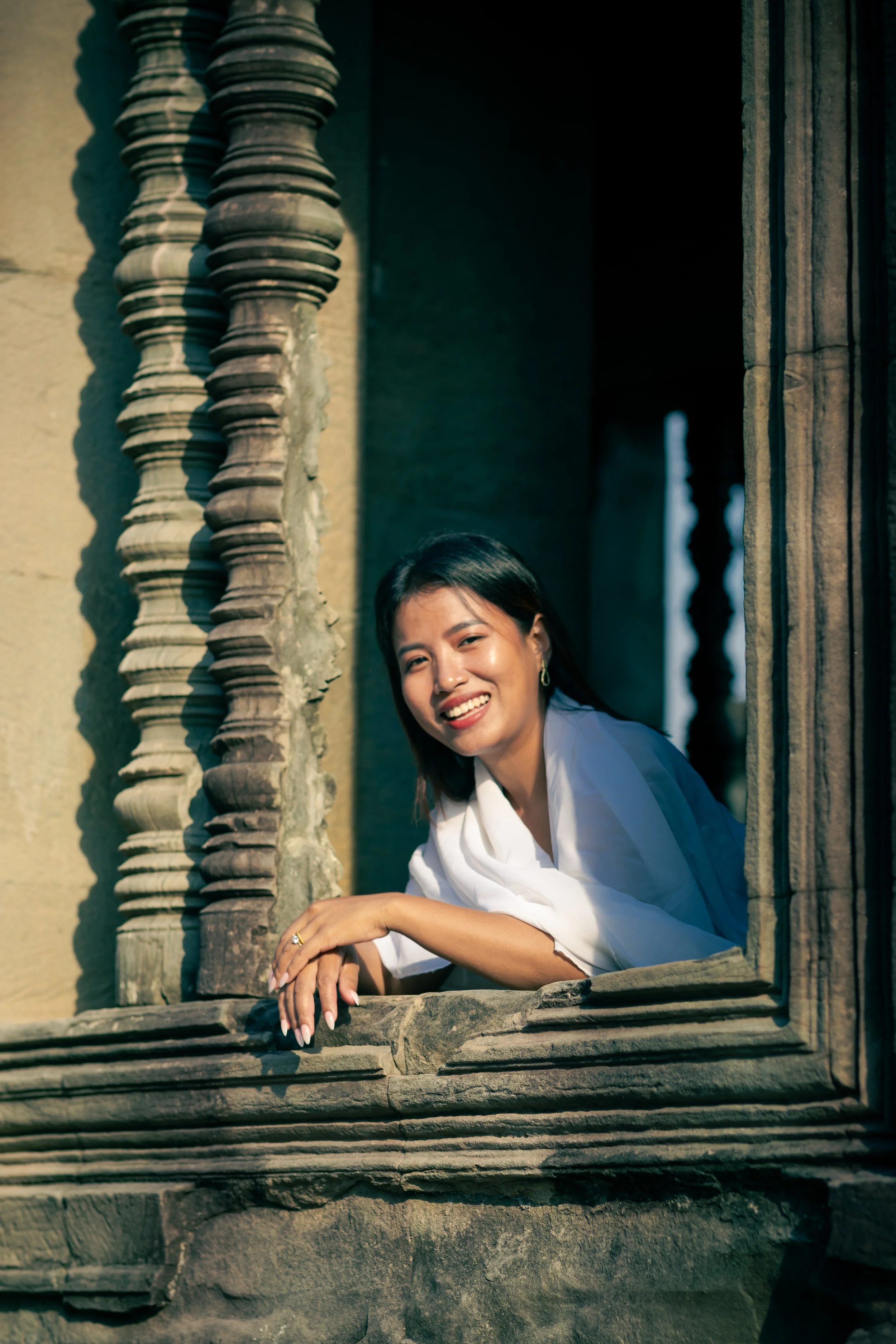 Woman smiling, leaning on stone window ledge with ornate columns at Angkor Wat in Siem Reap, Cambodia.