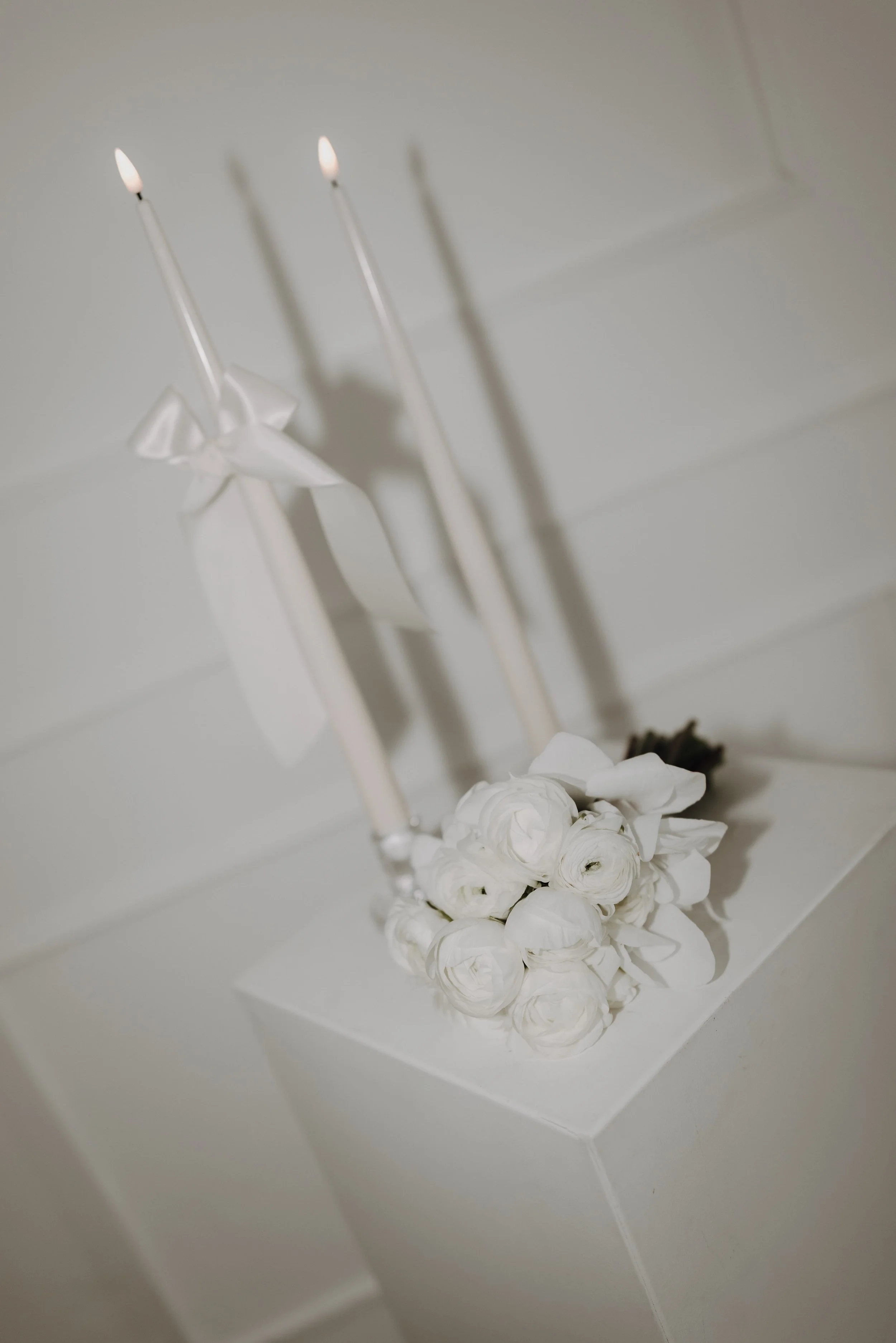 Weiße Rosen, zwei angezündete Kerzen und eine weiße Schleife liegen auf einer weißen Oberfläche.