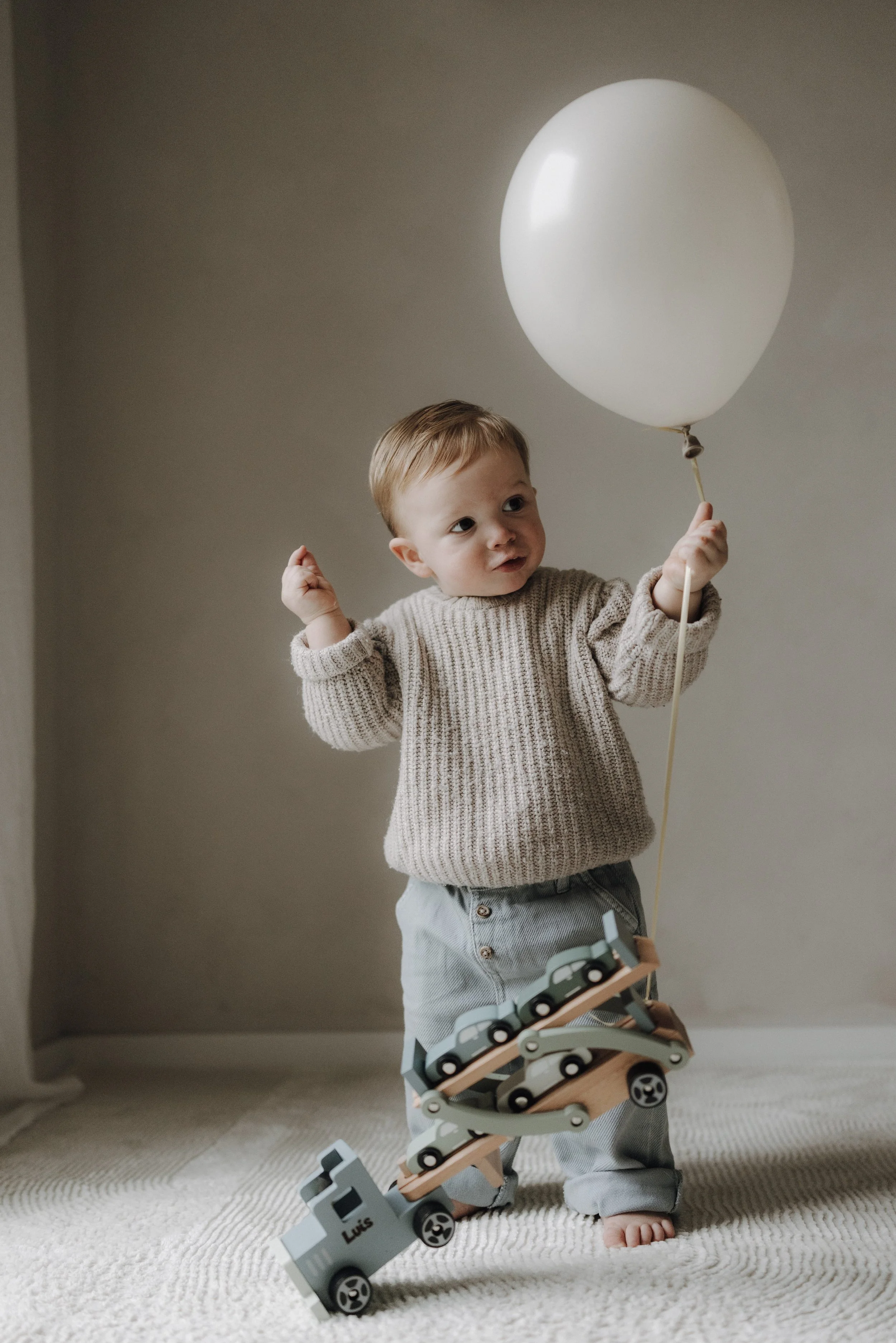 Ein kleines Kind hält einen weißen Luftballon in der Hand und spielt mit einem Spielzeuglaster aus Holz, das den Namen "Luis" trägt, auf einem Teppichboden in einem neutral gestrichenen Raum.