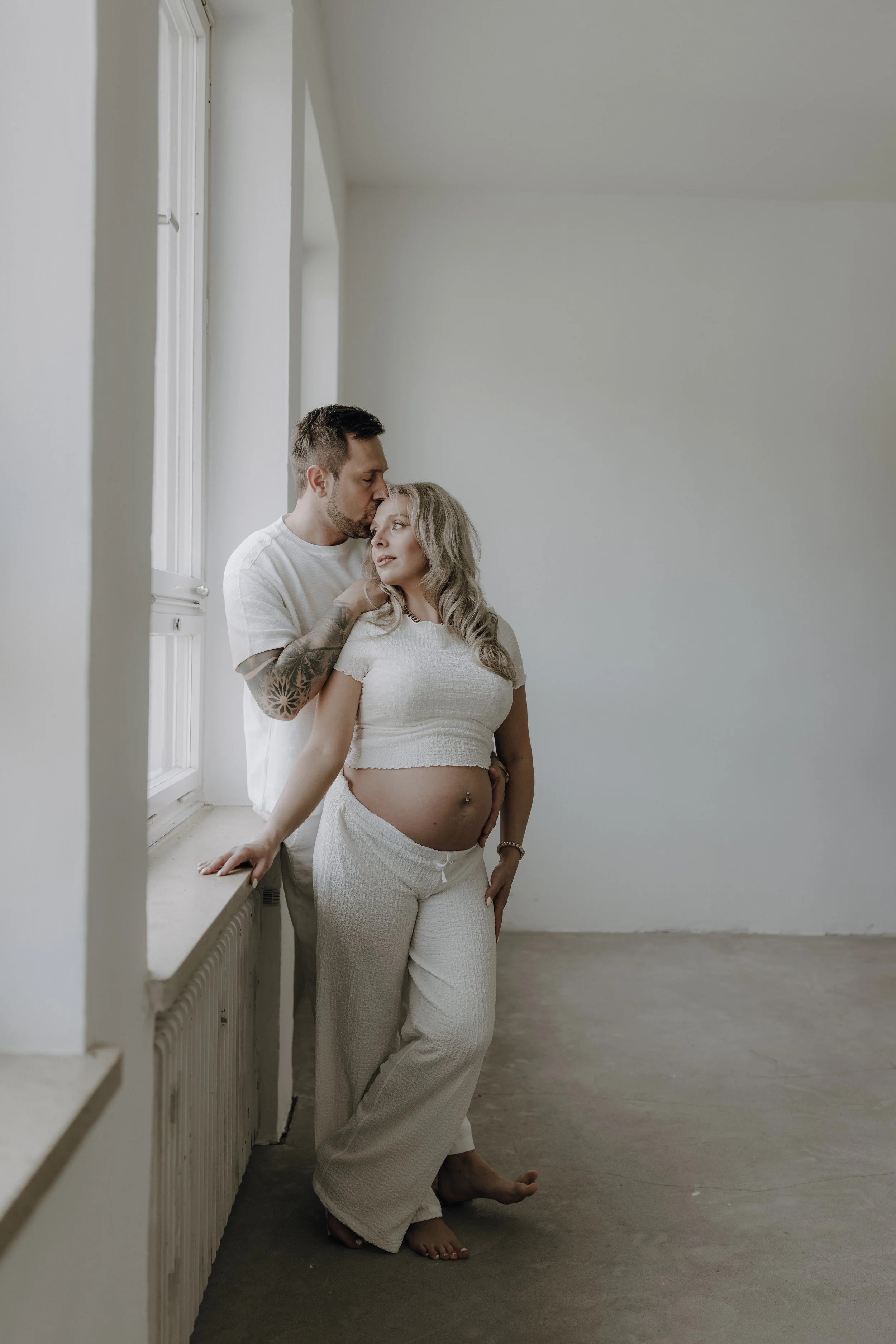 Ein schwangertes Paar in einem hellen, minimalistischen Raum, nahe am Fenster, bei einem liebevollen Moment.