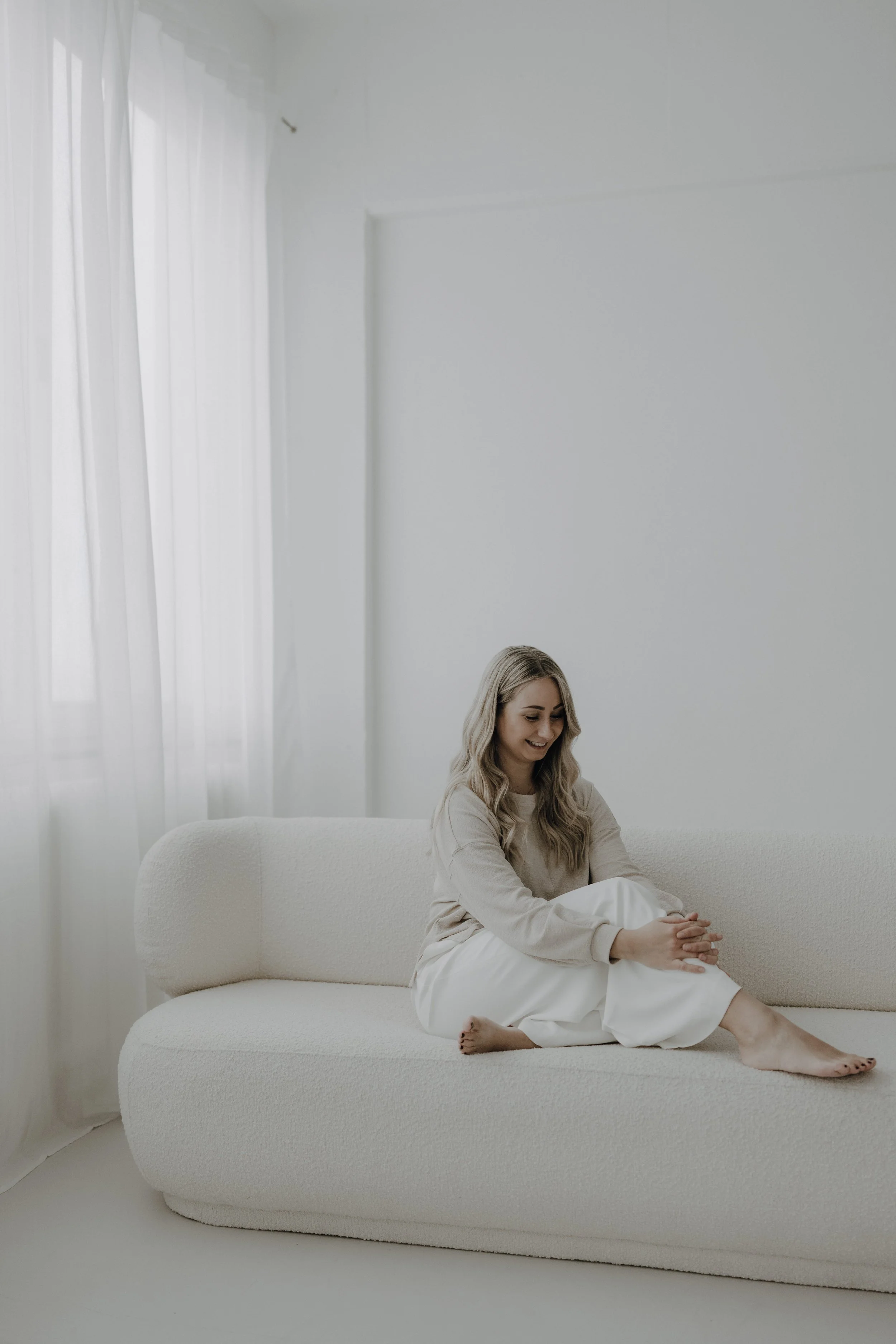 Eine junge Frau sitzt entspannt auf einer weißen Couch im hell erleuchteten, minimalistischen Raum mit weißen Wänden und Vorhängen.