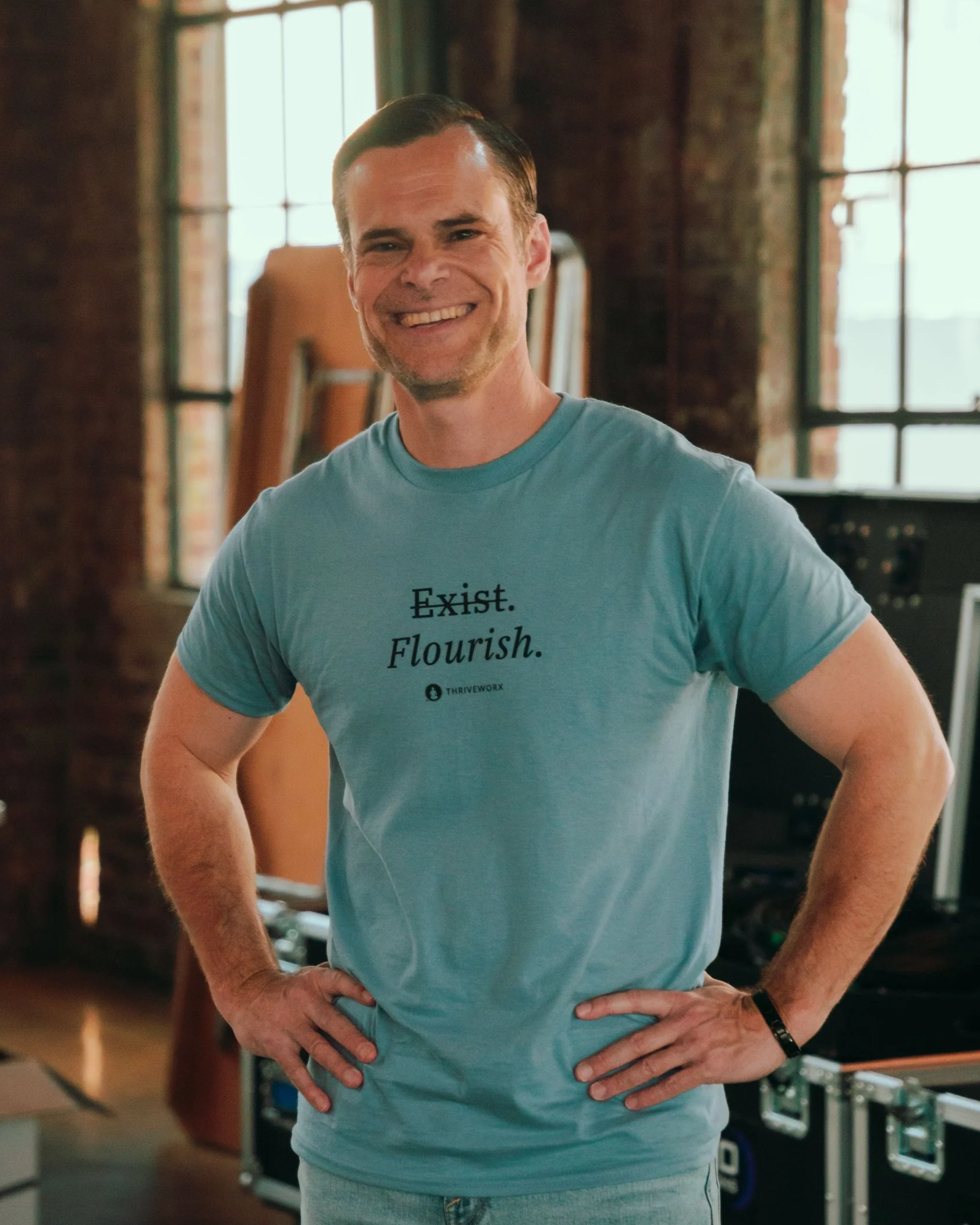 A smiling man standing indoors with large windows behind him, wearing a blue T-shirt with the words 'Fight. Exist. Flourish.' and gray pants, with his hands on his hips.