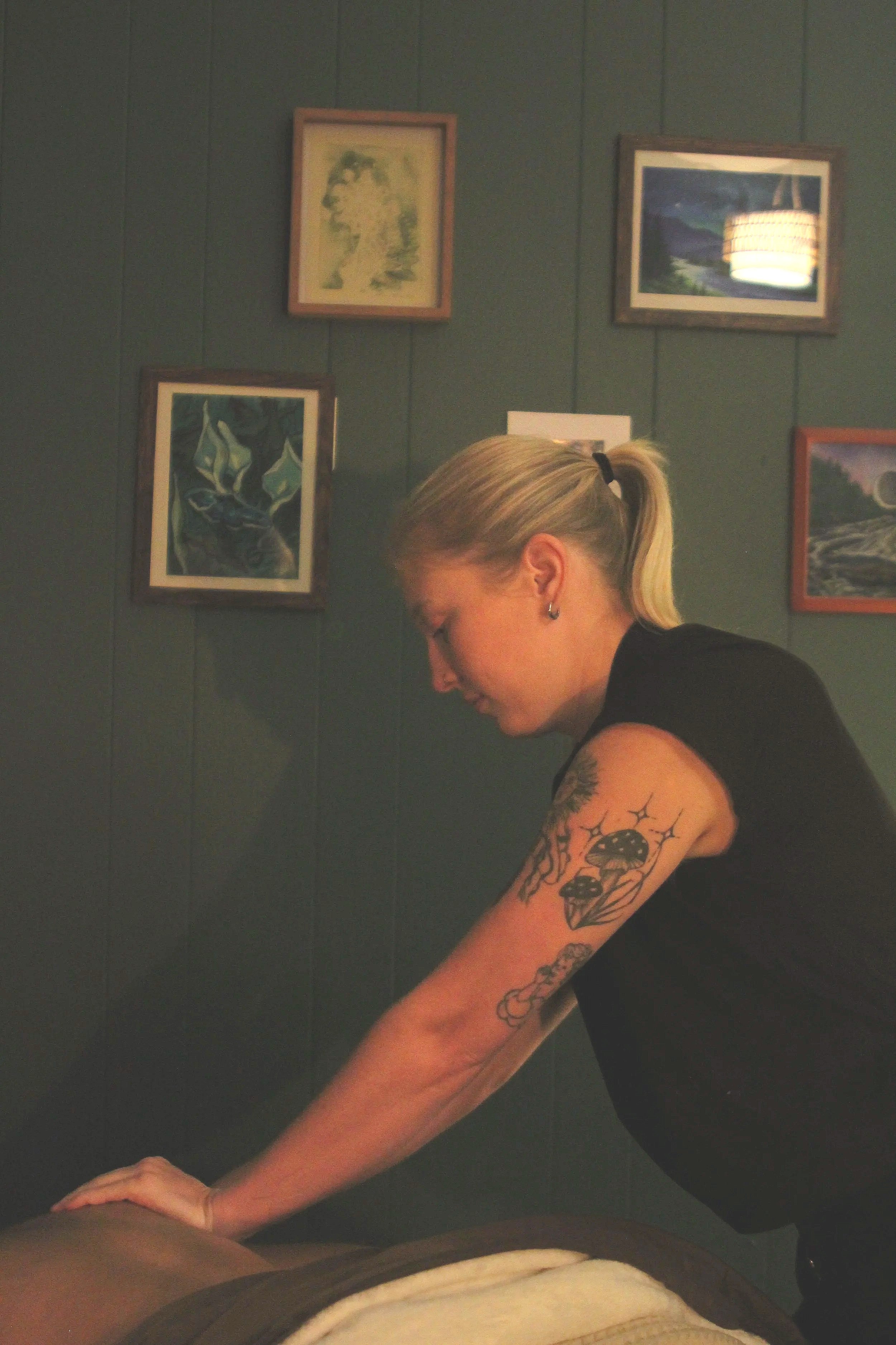 A woman with blonde hair tied back in a ponytail and tattoos on her arm is giving a massage in a room with dark green walls and framed artwork. The artwork includes a  landscapeand abstract images. There is a hanging lampshade reflecting on the wall.