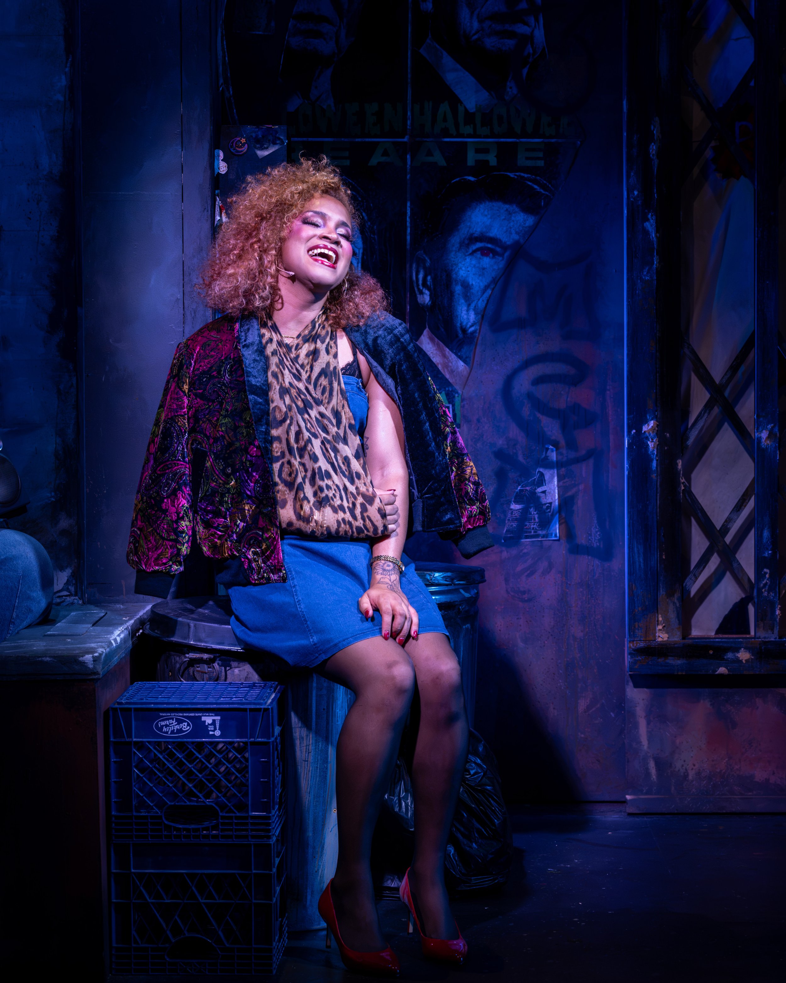 Audrey sits outside the flower shop, gazing upward under blue stage lighting in Little Shop of Horrors.