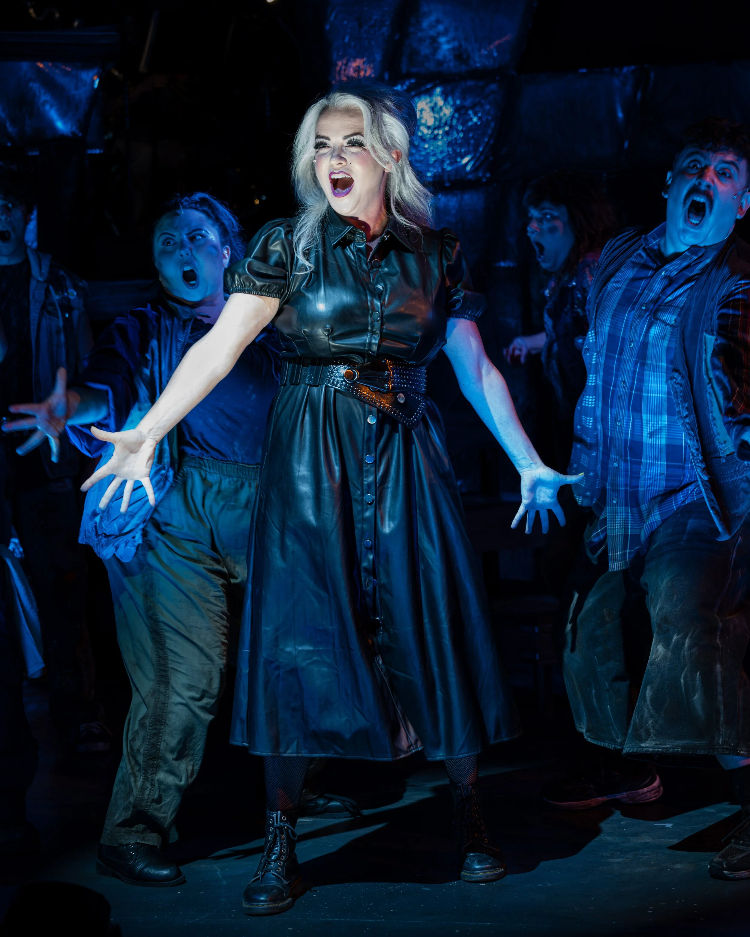 Penelope Pennywise sings with her arms outstretched as the ensemble surrounds her under vivid blue stage lighting in Urinetown.