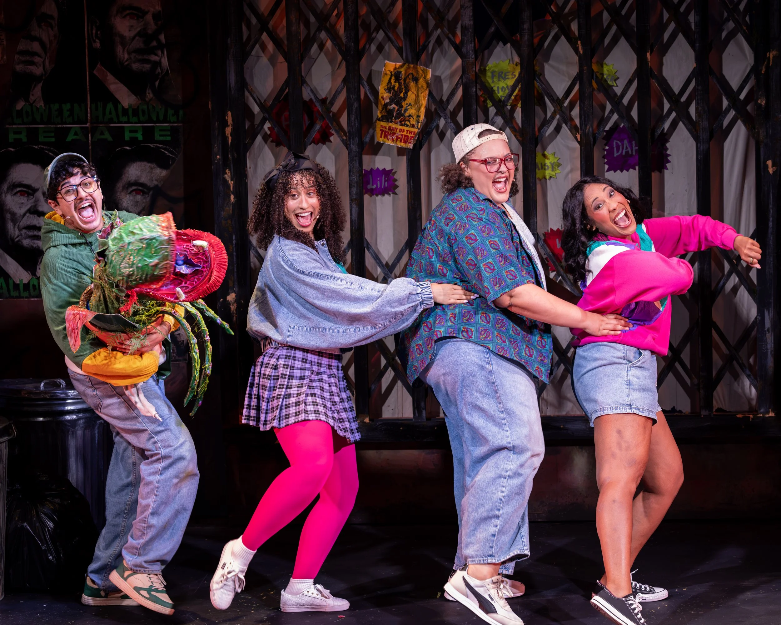 Seymour carries a small Audrey II puppet while three performers dance beside him in colorful costumes in Little Shop of Horrors.