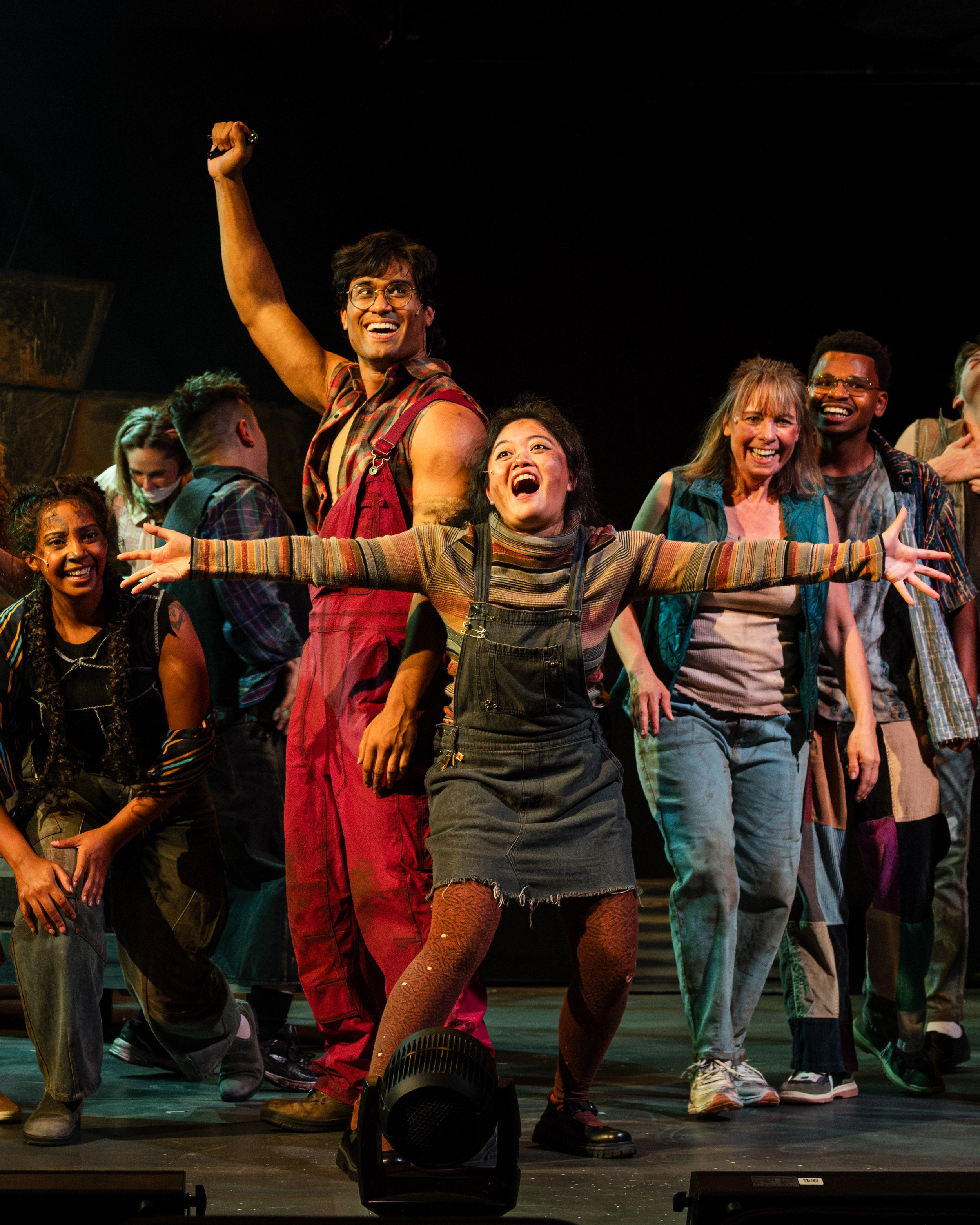 Little Sally stretches her arms wide as Bobby Strong raises a fist and the ensemble celebrates behind them in Urinetown.