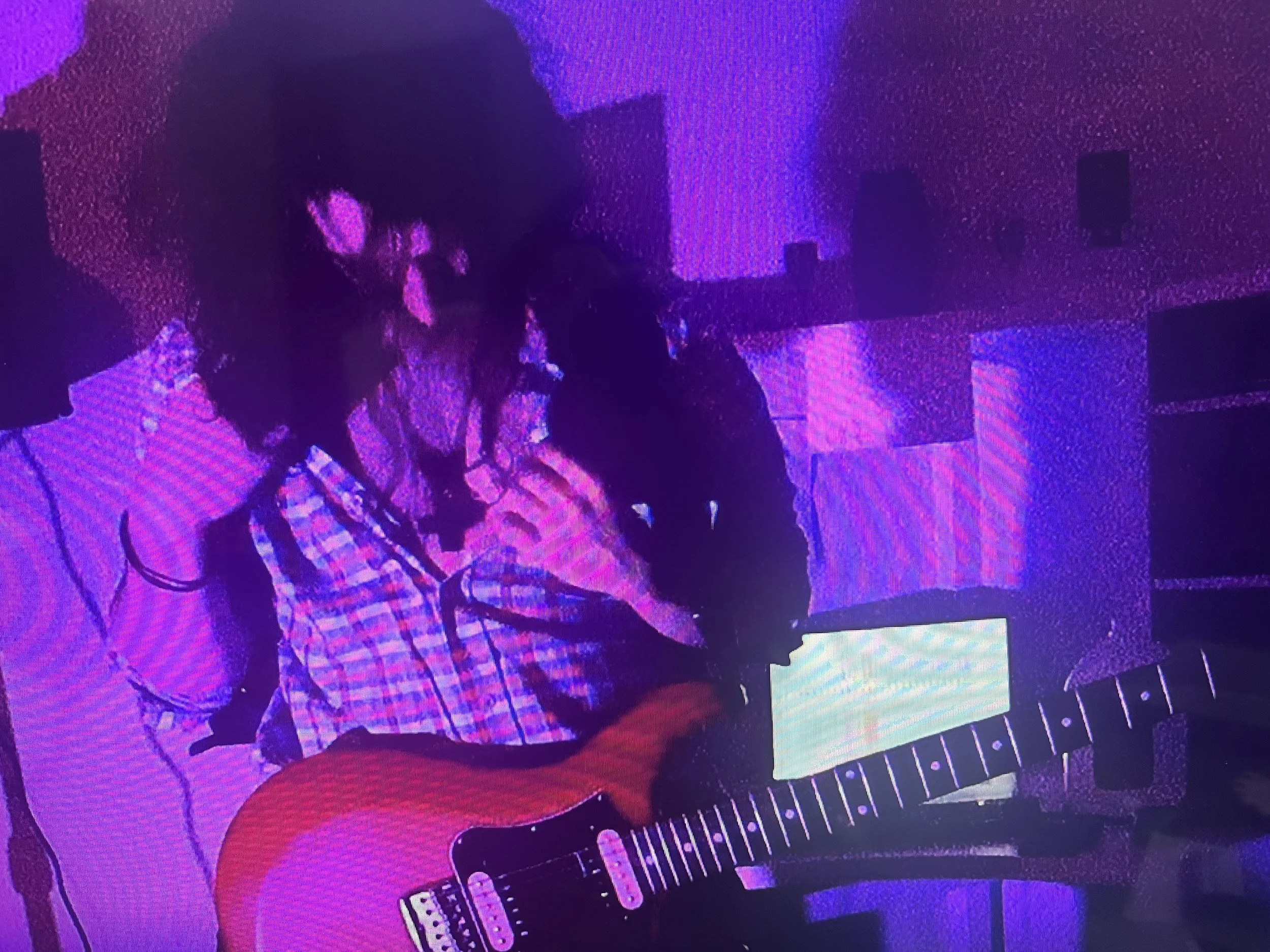 Person with long, dark, and flowing hair with a black cross necklace and a guitar standing in a pink and blue lit room in front of white desk and a computer screen.