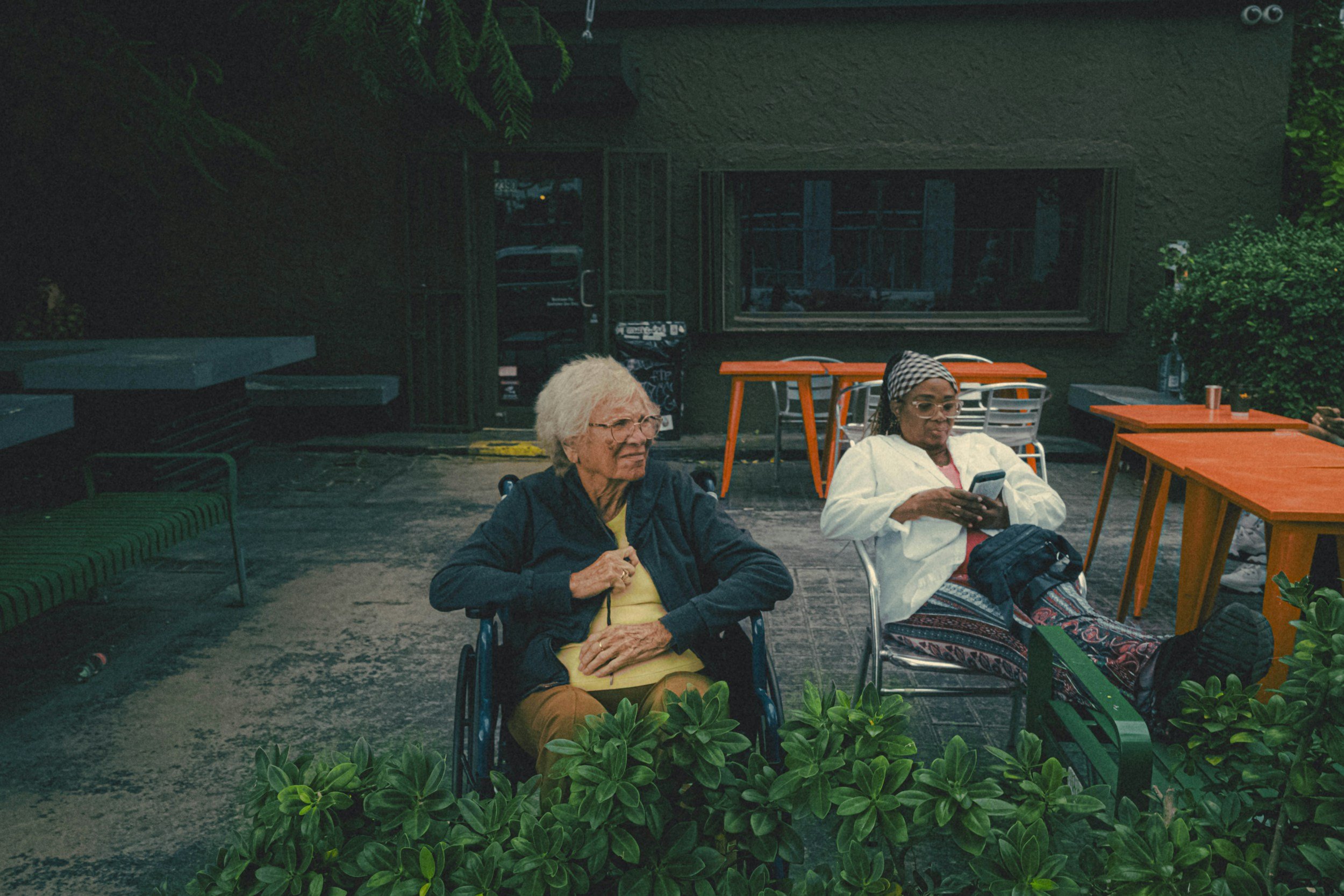 elderly woman in a wheelchair and her caretaker sitting in a chair nearby