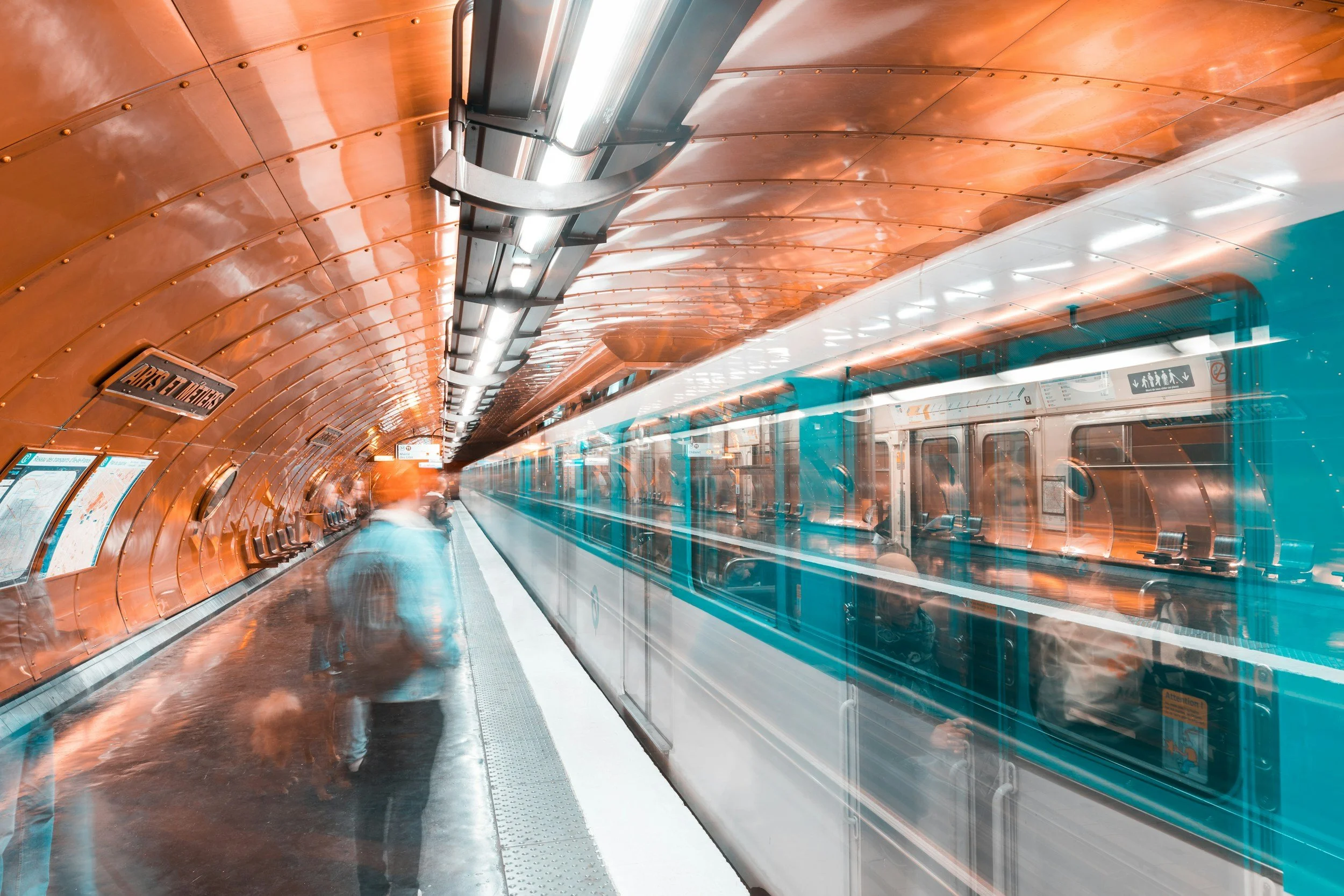 Underground subway station with a moving train, orange curved ceiling, and blurred passengers on the platform.