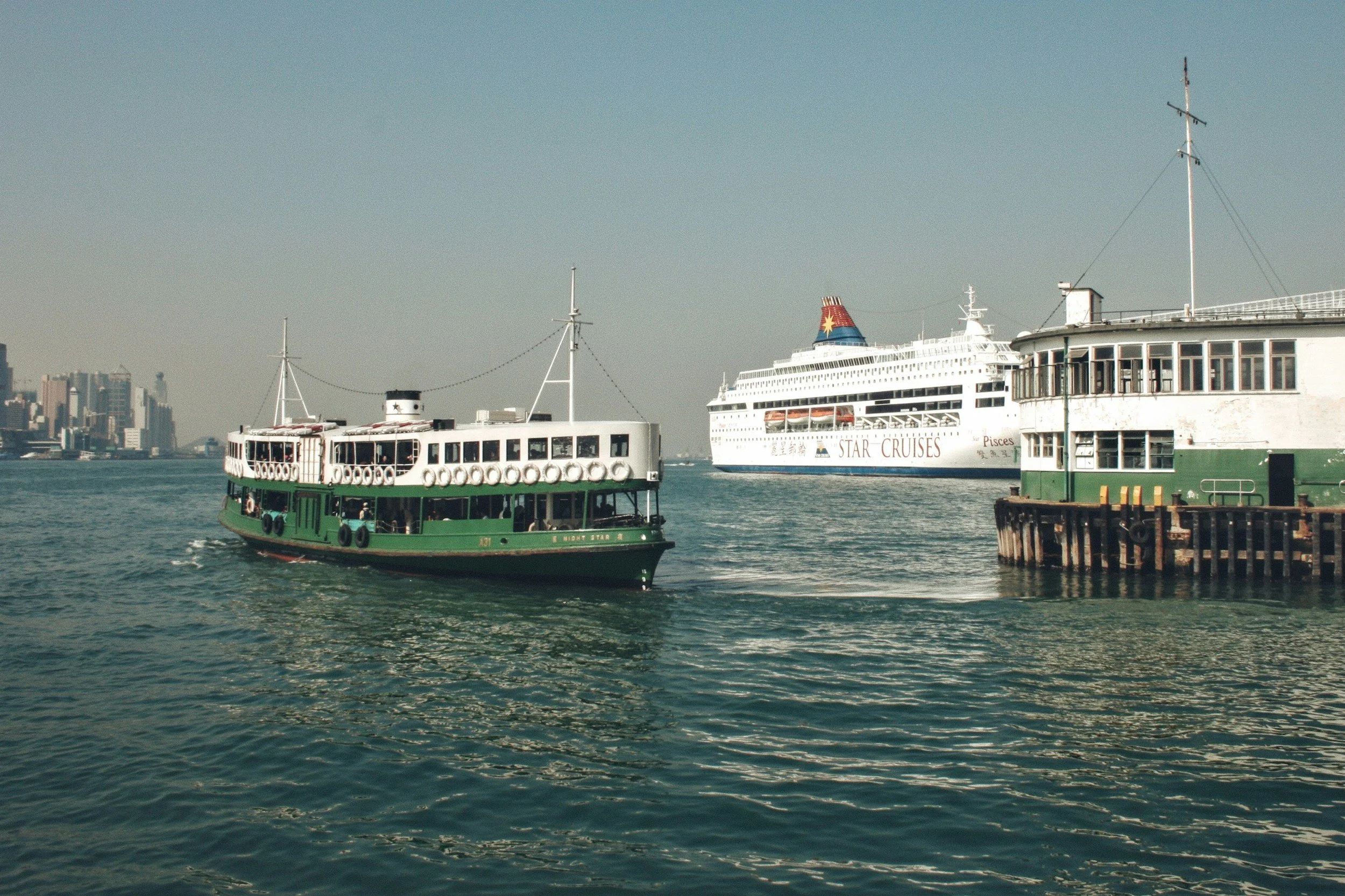 Two boats on the water with a city skyline in the background. One boat is green and white, the other is white with a colorful funnel.