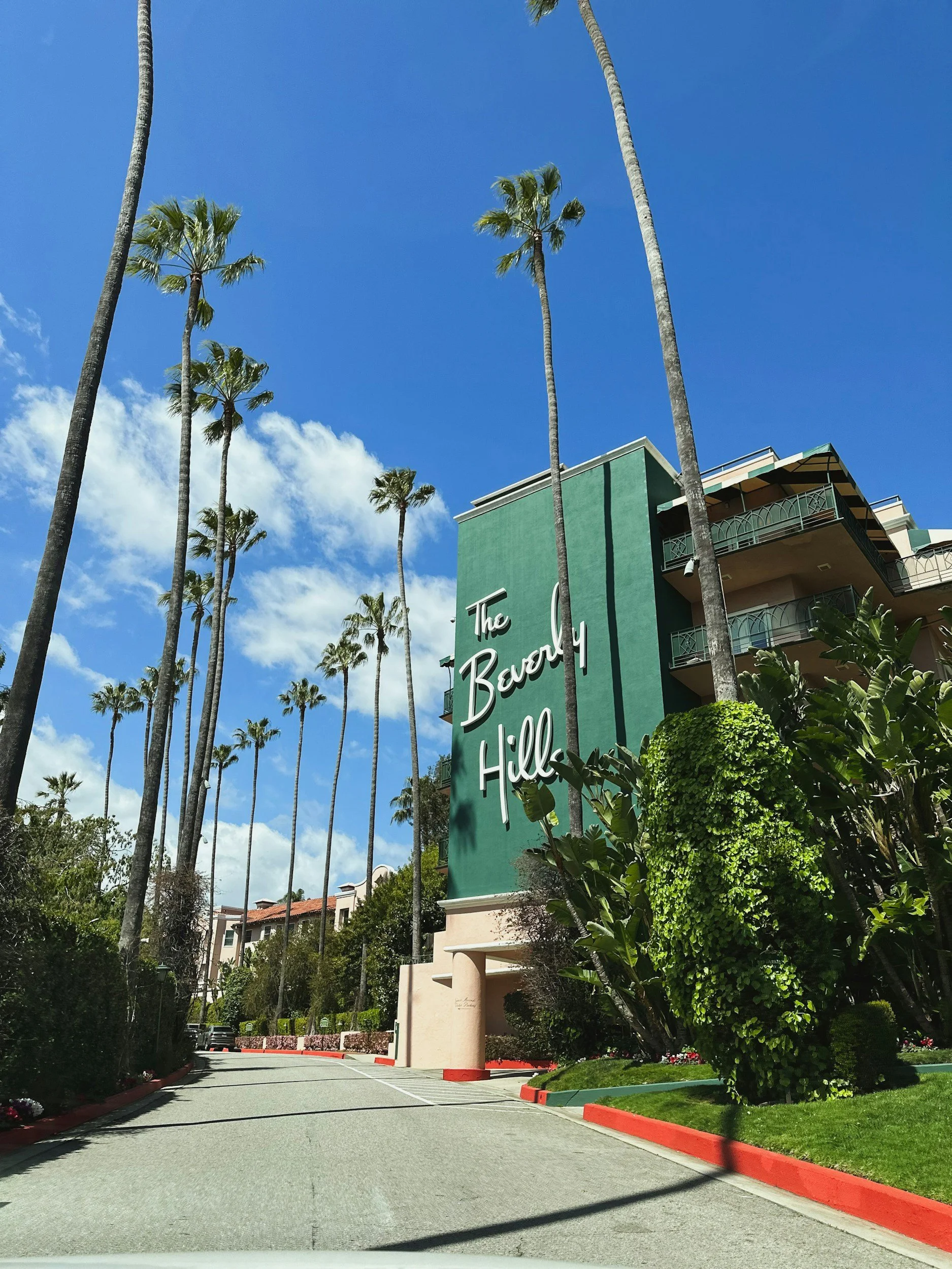 A tall green building with white cursive text reading 'The Beverly Hill' on it, surrounded by palm trees and greenery under a partly cloudy blue sky.