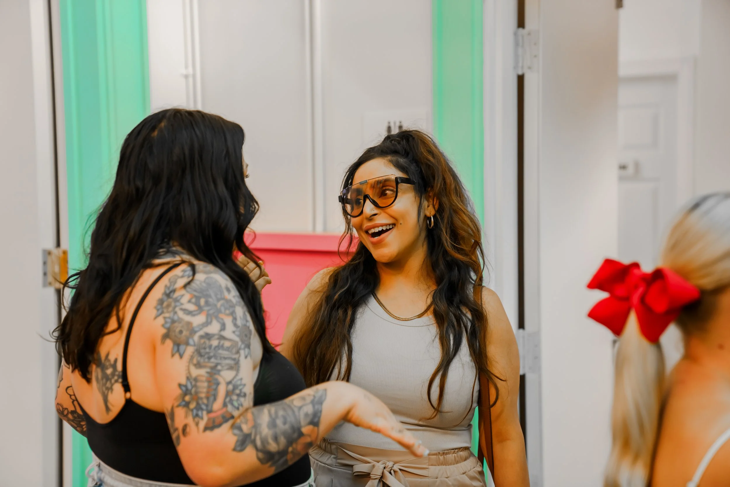 Three women talking and smiling indoors at a social gathering. One woman with tattoos on her arms and long dark hair facing the camera, another woman with curly hair, wearing sunglasses and a beige top, smiling and looking at the tattooed woman, and