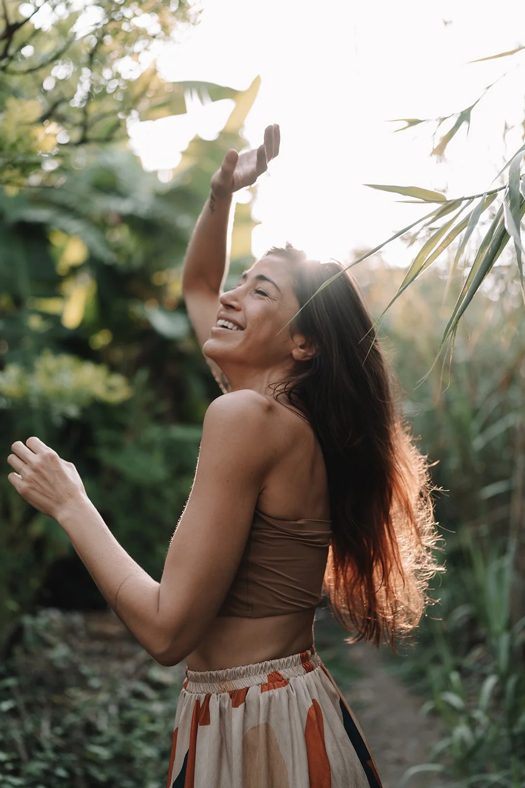 A woman with long hair smiling and dancing in a lush, green outdoor setting with sunlight filtering through trees.