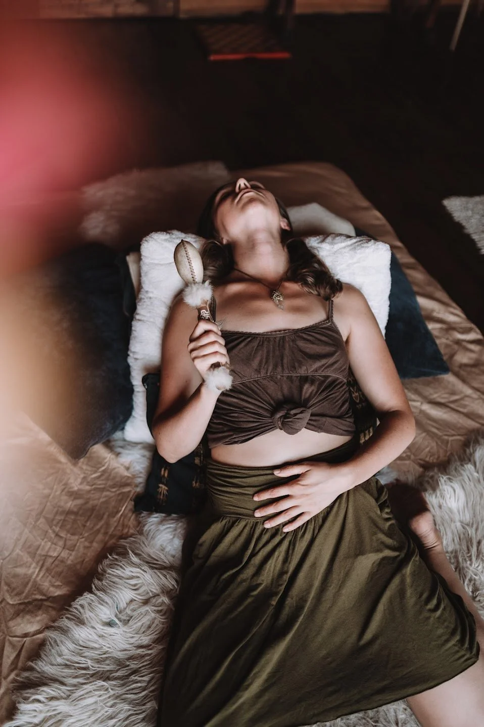 A woman lying on a bed with her eyes closed, holding a decorative object resembling a scepter, wearing a brown tank top and a green skirt.